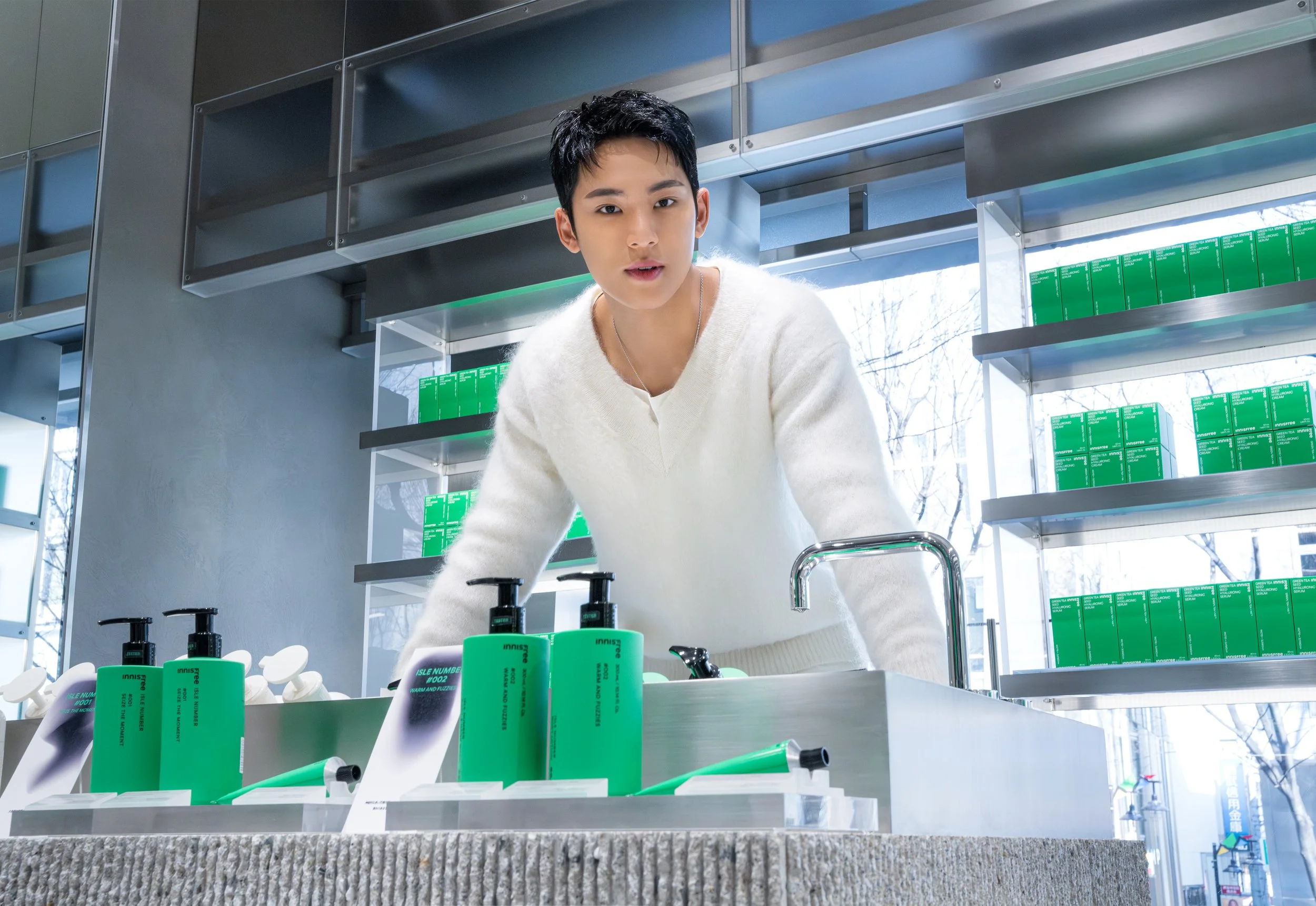 Young man in a white sweater looking over a display of green bottles with pumps in a modern store with glass shelves and large windows