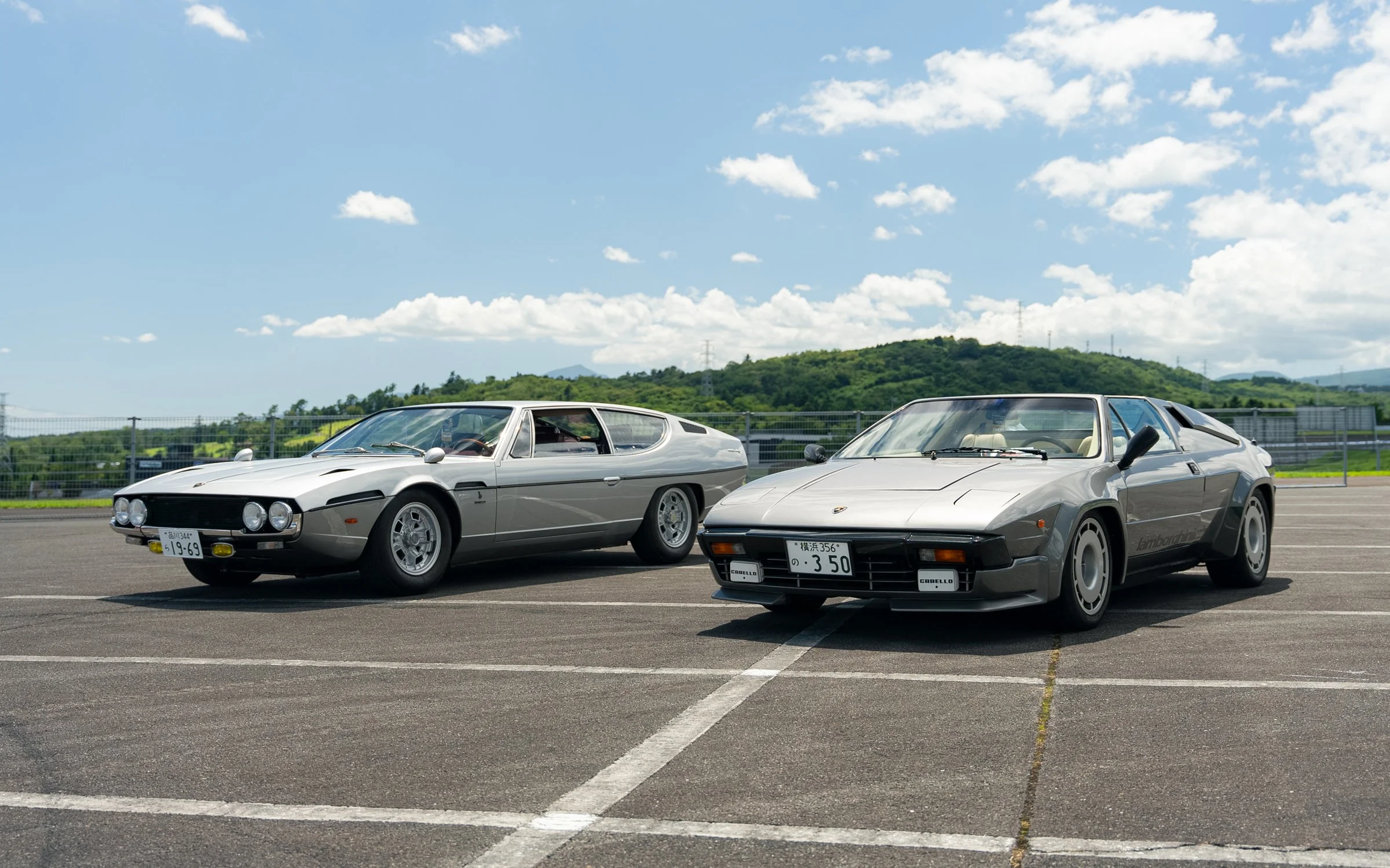 Two vintage sports cars, a Lamborghini and a Ferrari, parked on an empty parking lot with a scenic green mountain and blue sky background.