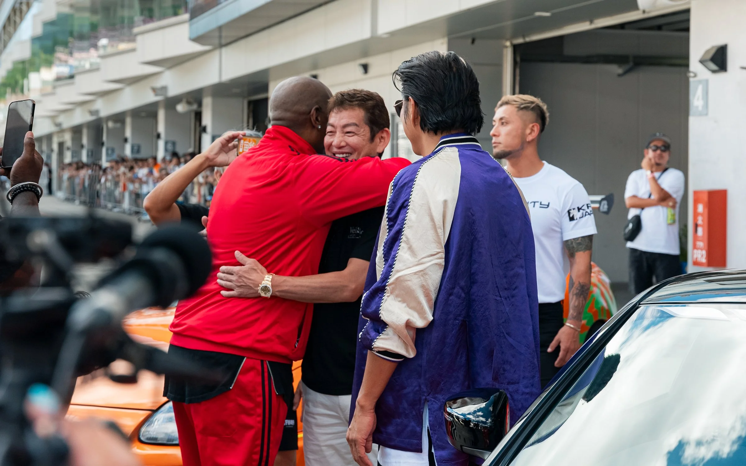 Formula 1 driver celebrating with group, hugging and smiling, at race event with fans in background.