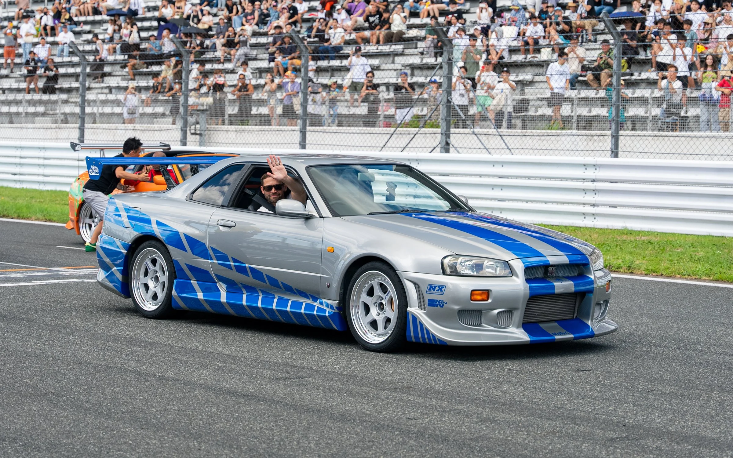 A silver sports car with blue racing stripes and graphics is on a racetrack, with a man waving from the passenger side window. In the background, there is a crowd of spectators seated in bleachers and a person working on a car.