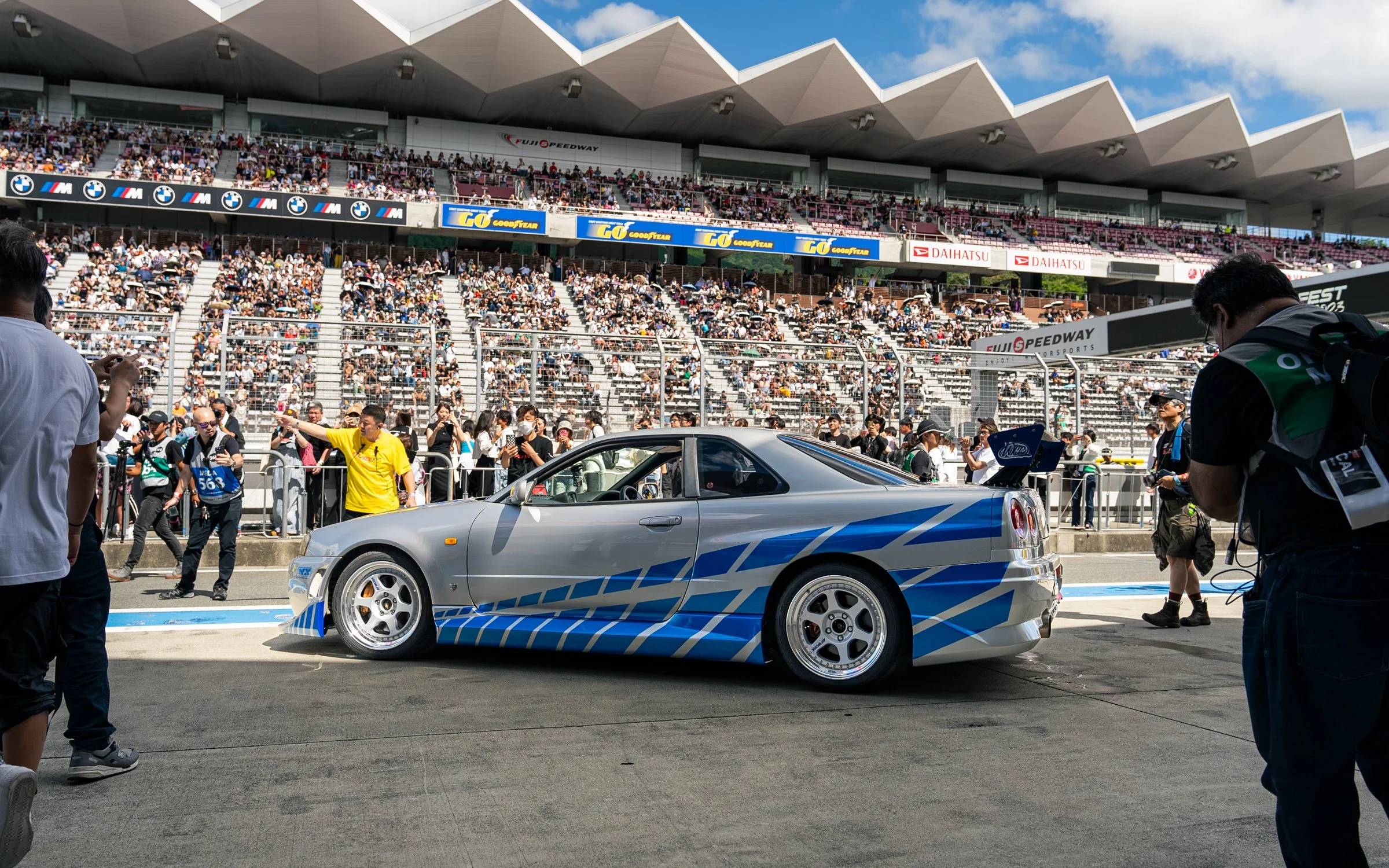 A silver race car with blue stripes parked at Fuji Speedway during a racing event, surrounded by attendees and photographers.