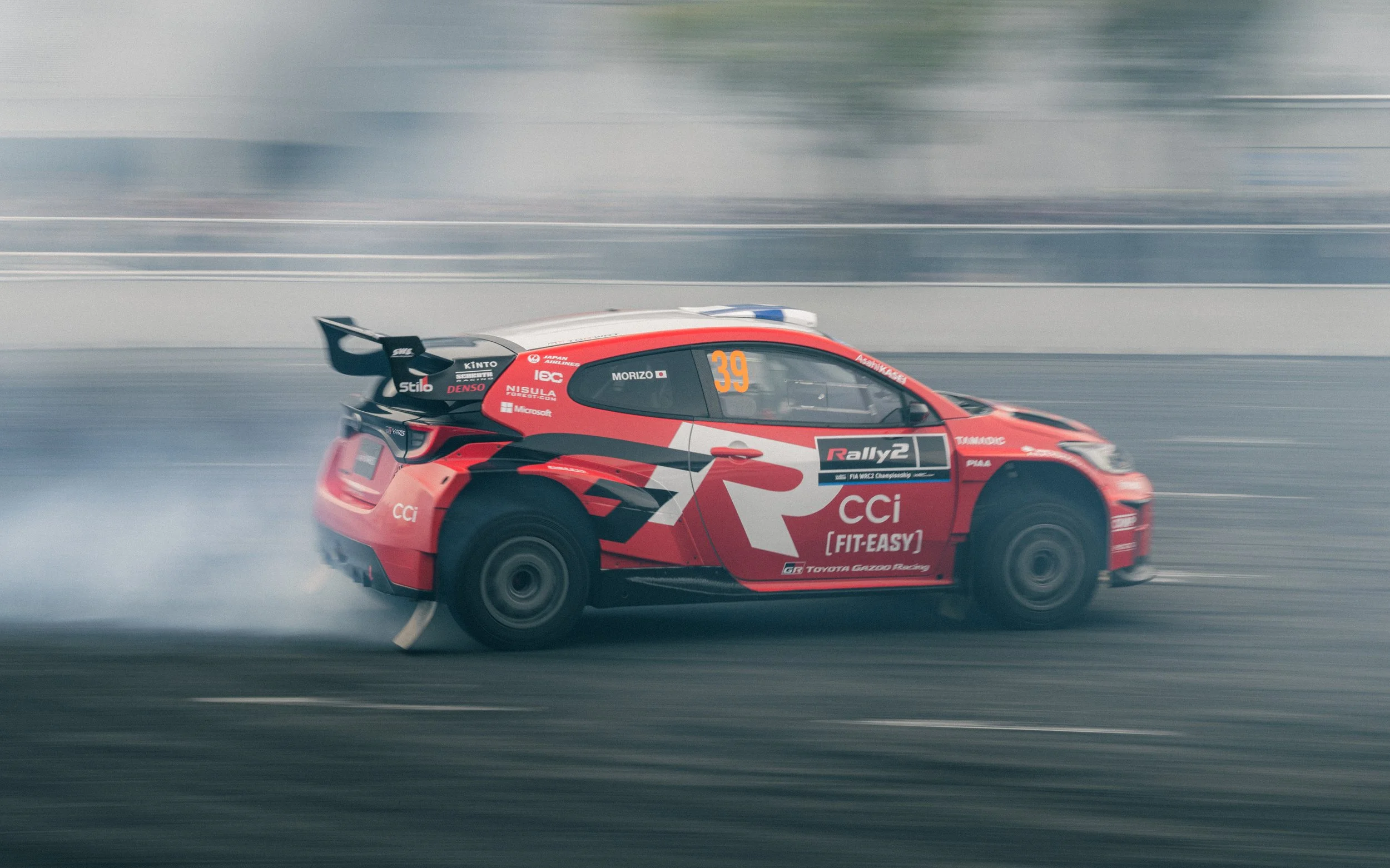A red and black rally car racing on a track, leaving tire smoke behind as it drifts.