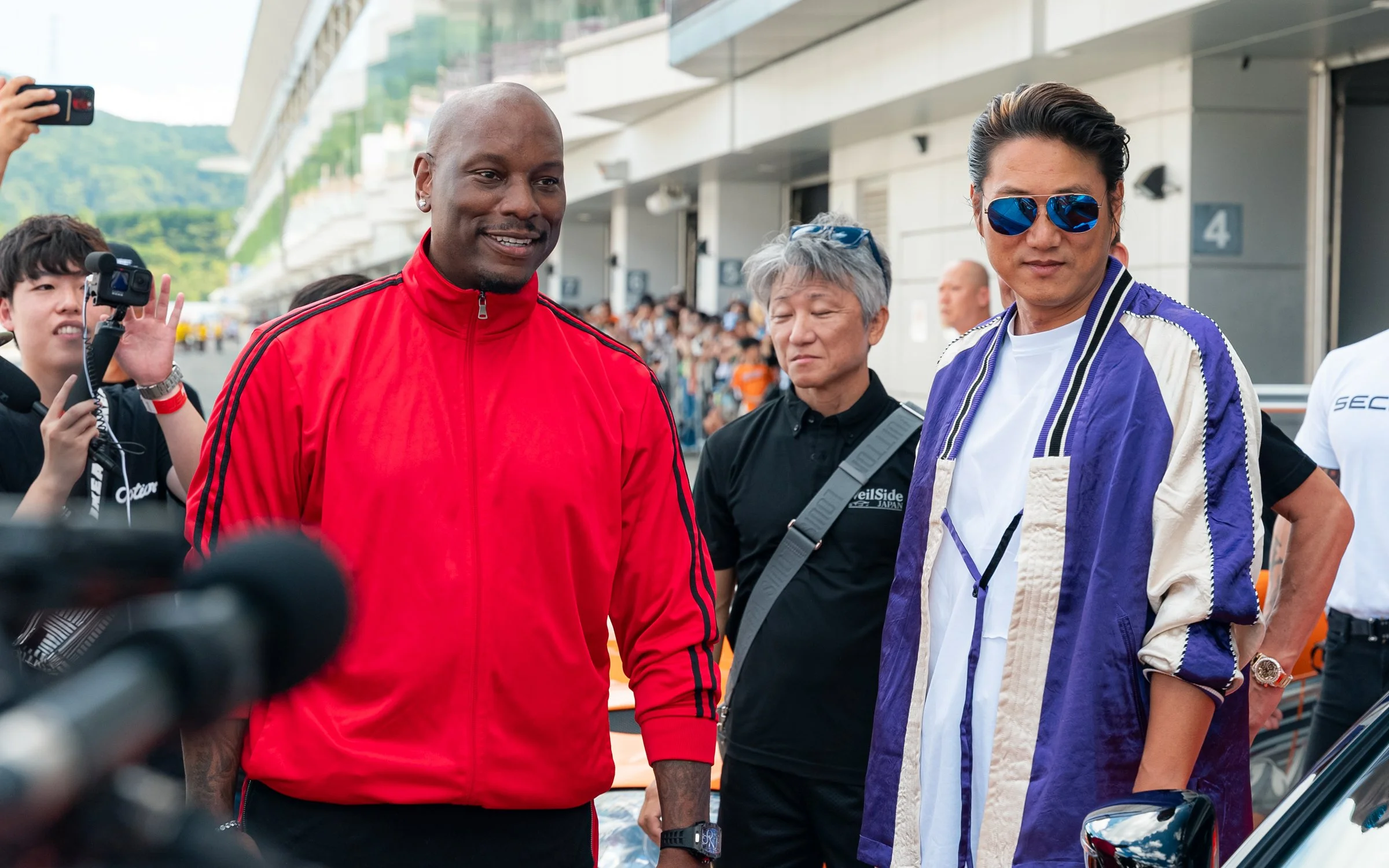 Two men are standing outdoors at a racing event, one in a red jacket and the other in a purple and white jacket with blue sunglasses, while several people, some with cameras, are in the background.