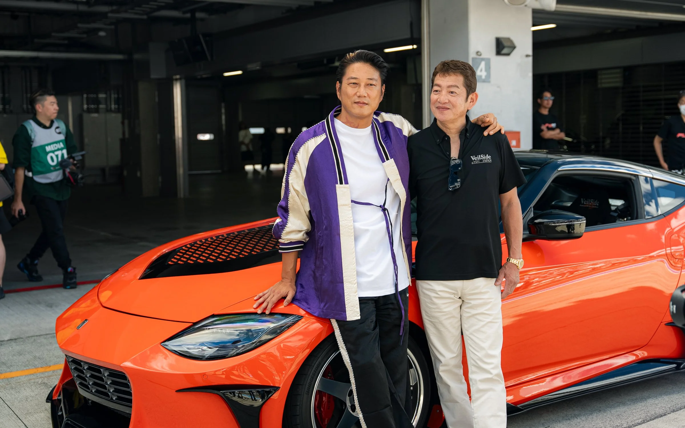 Two men standing next to an orange sports car, with one resting his hand on the car. They are smiling and posing for the photo in a garage or pit area, with other people in the background.