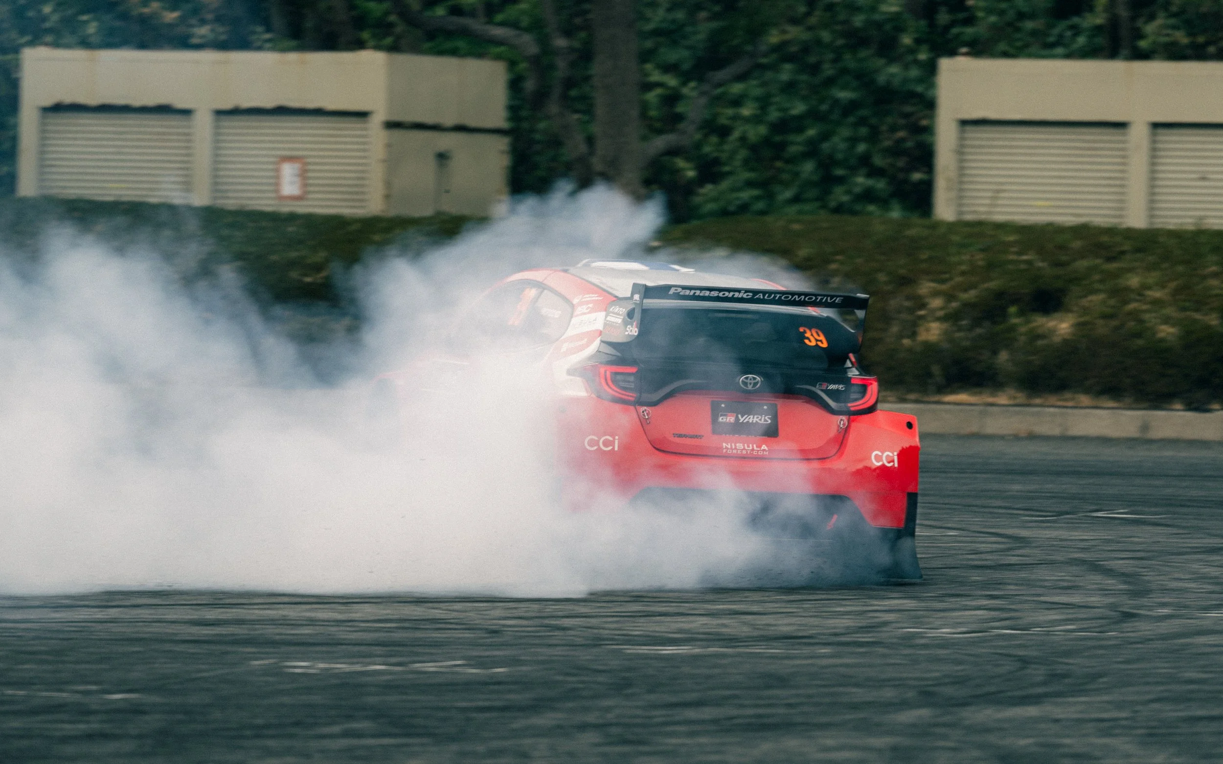 A red Toyota racing car, number 39, drifting on a race track with smoke billowing from its tires, surrounded by greenery and some buildings in the background.