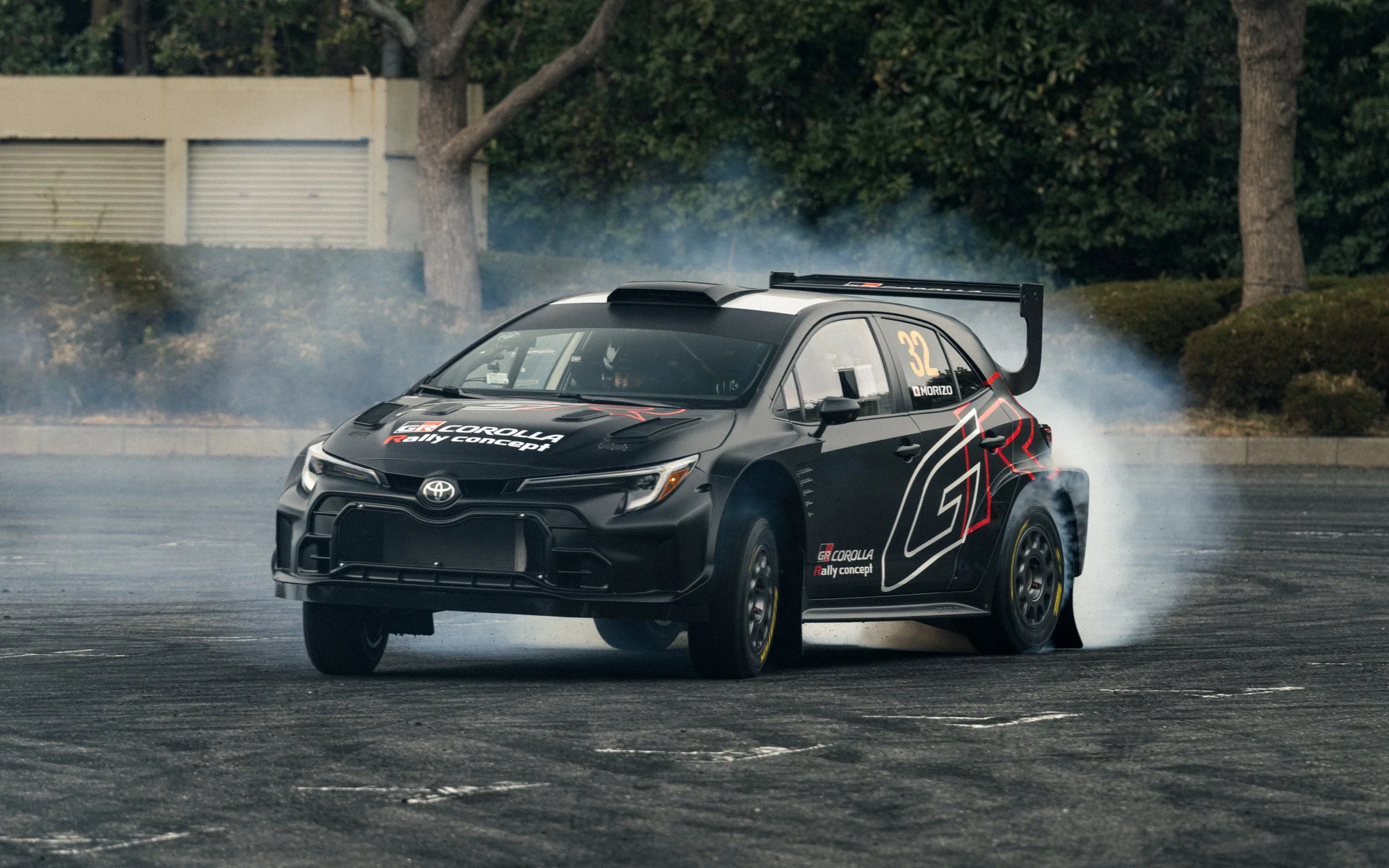 A black Toyota GR Corolla rally car drifting on a paved surface, creating smoke from the tires, with trees and a fence in the background.