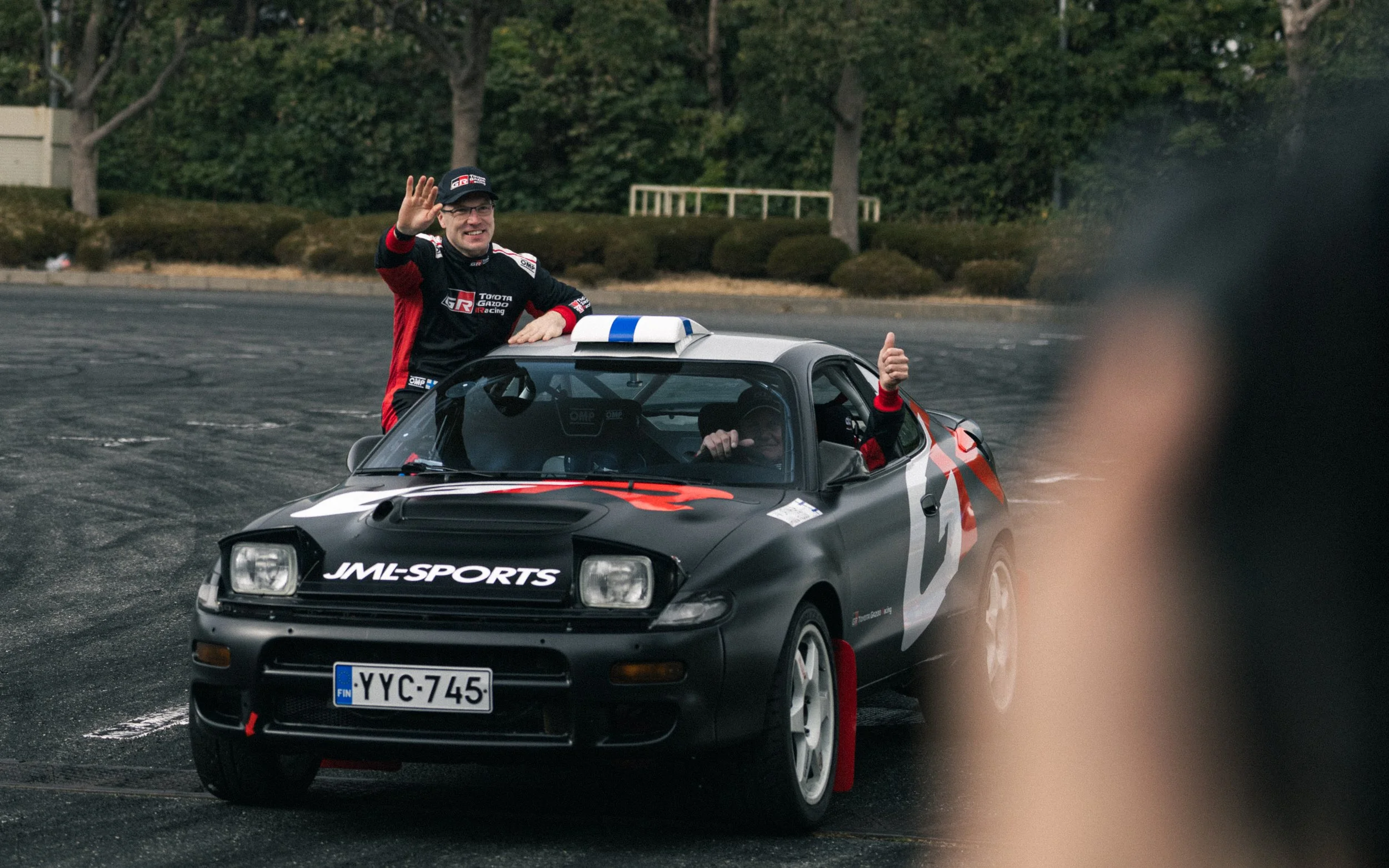 A race car driver and a passenger celebrating in a rally car with Finnish license plates, giving thumbs up and waving during a race or event, with trees and a road in the background.