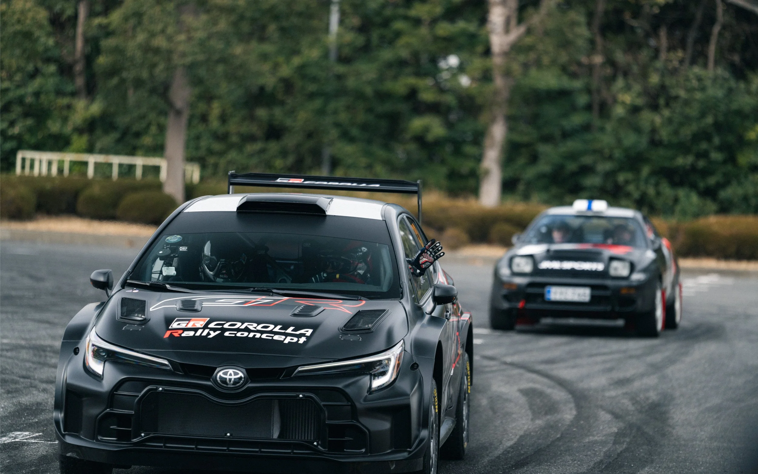 Two Toyota rally cars on a racetrack, racing with one another. The car in front is black with 'GR Corolla Rally Concept' written on the hood. The background shows a wooded area with trees.