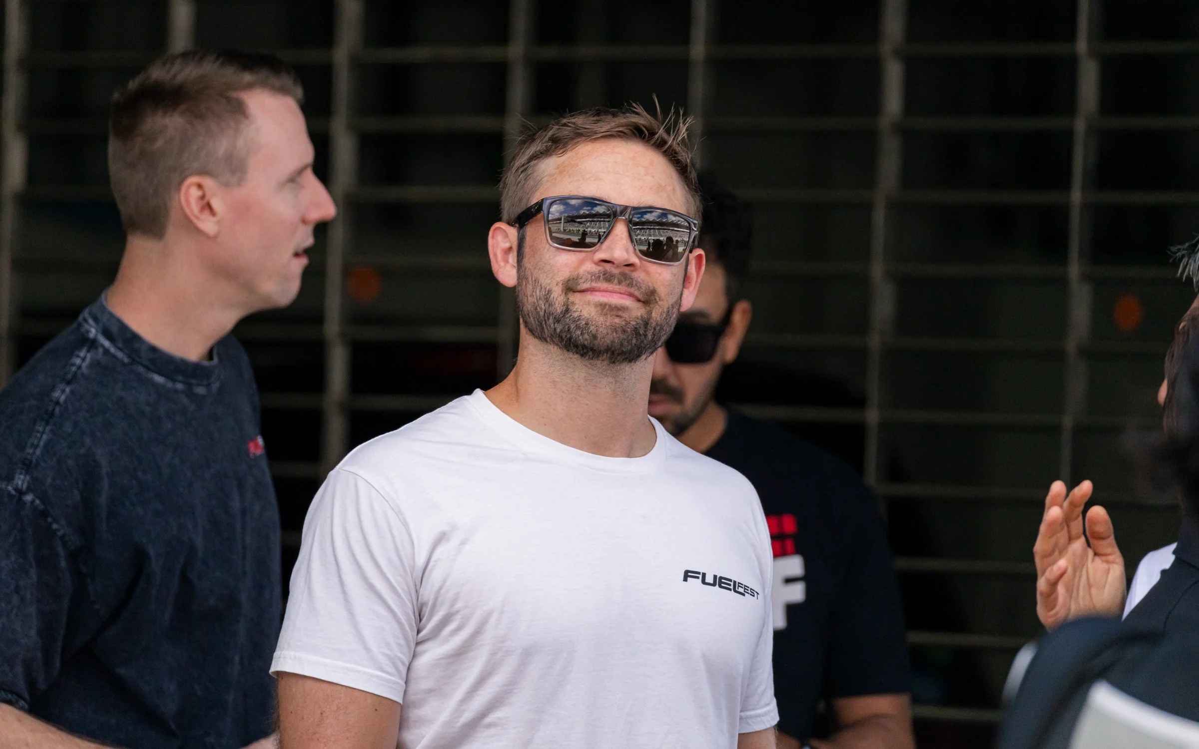 A man wearing sunglasses and a white T-shirt with 'FUEL FEST' on it, smiling while surrounded by other people in a discussion.