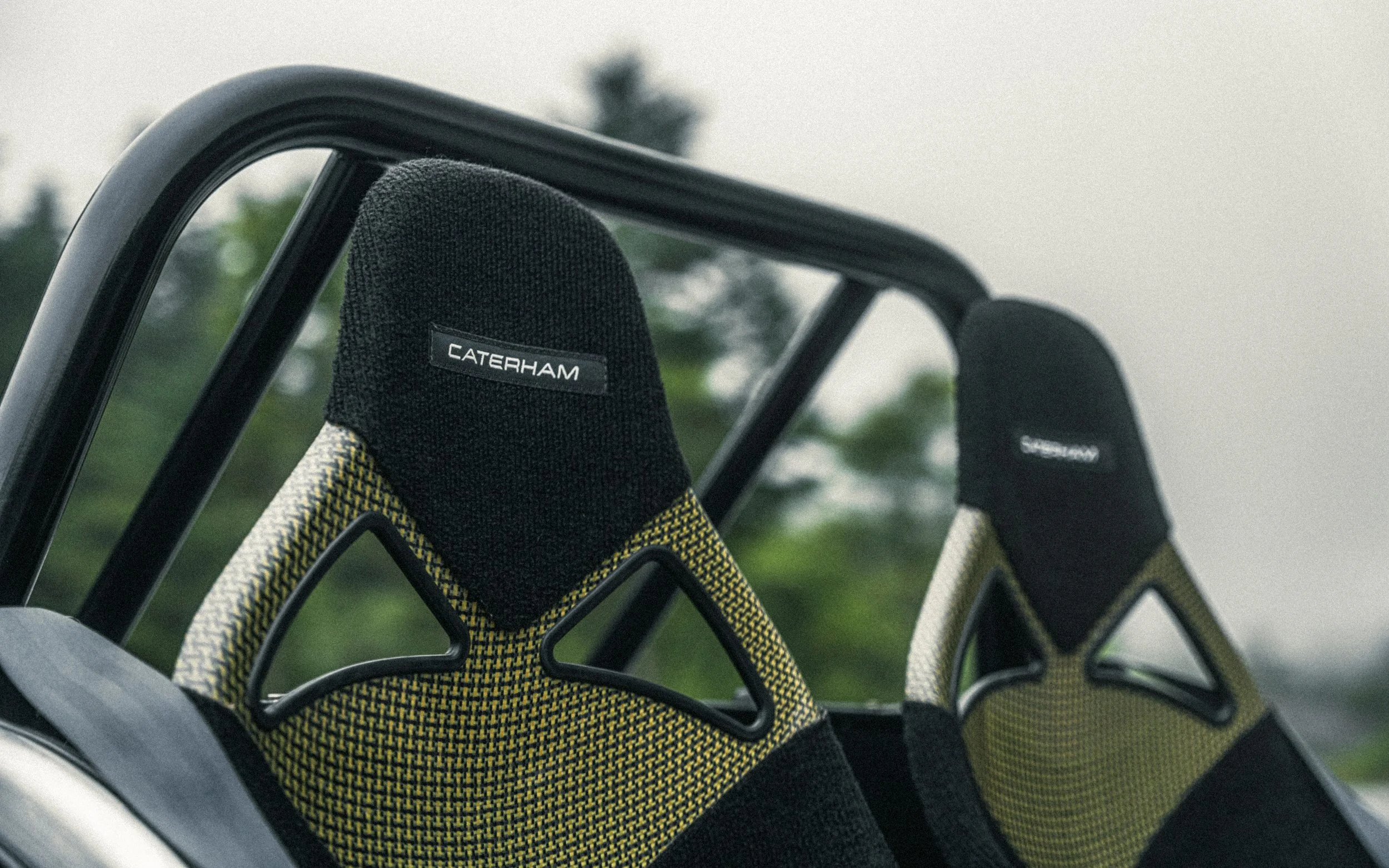 Two Caterham racing seats with headrests in an open vehicle, with a blurred outdoor background.