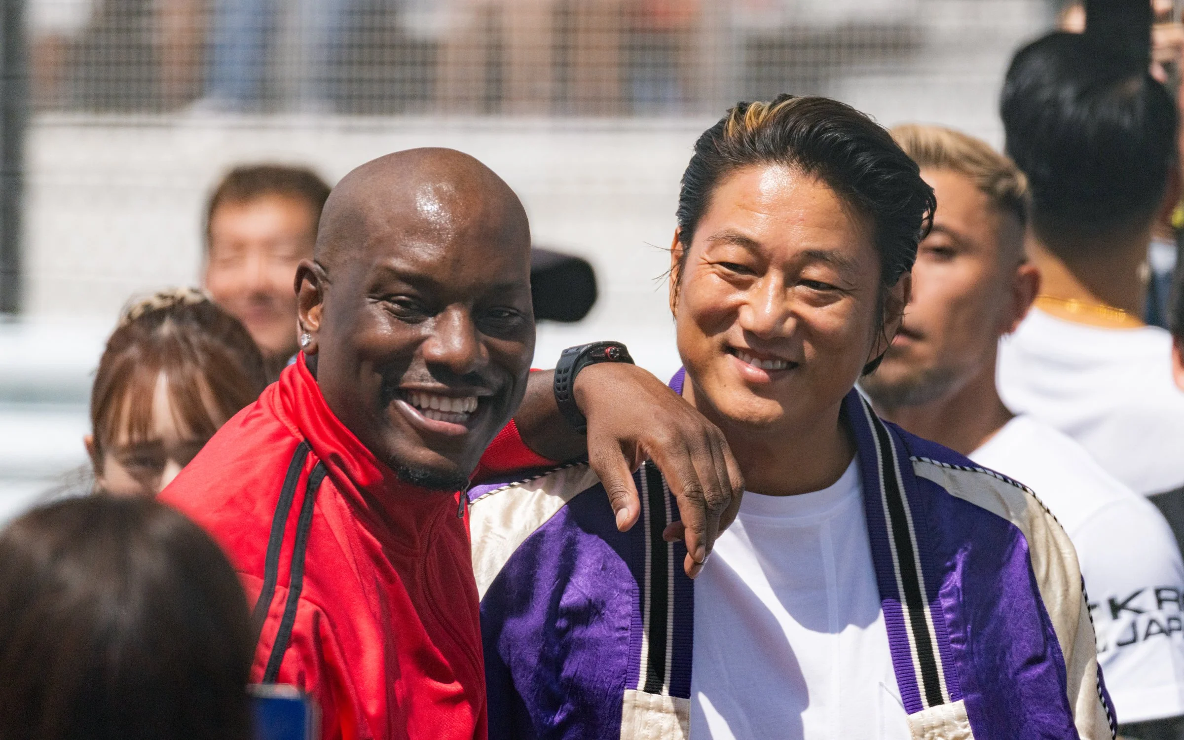 Two men smiling, one with his arm around the other's shoulder, at a social gathering outdoors with people in the background.
