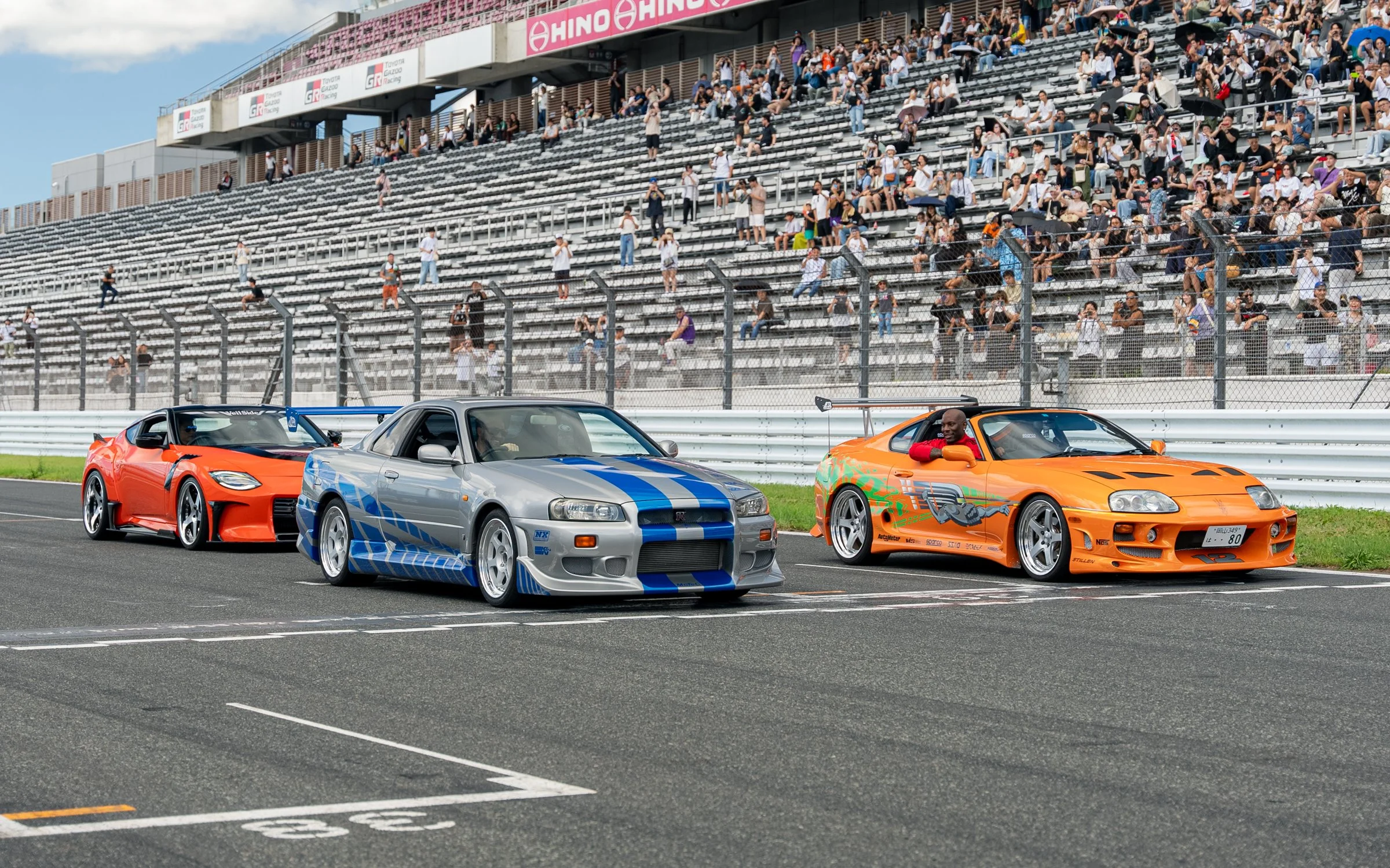 Three race cars parked at the starting line of a racing track, with a crowd of spectators in the grandstands in the background.
