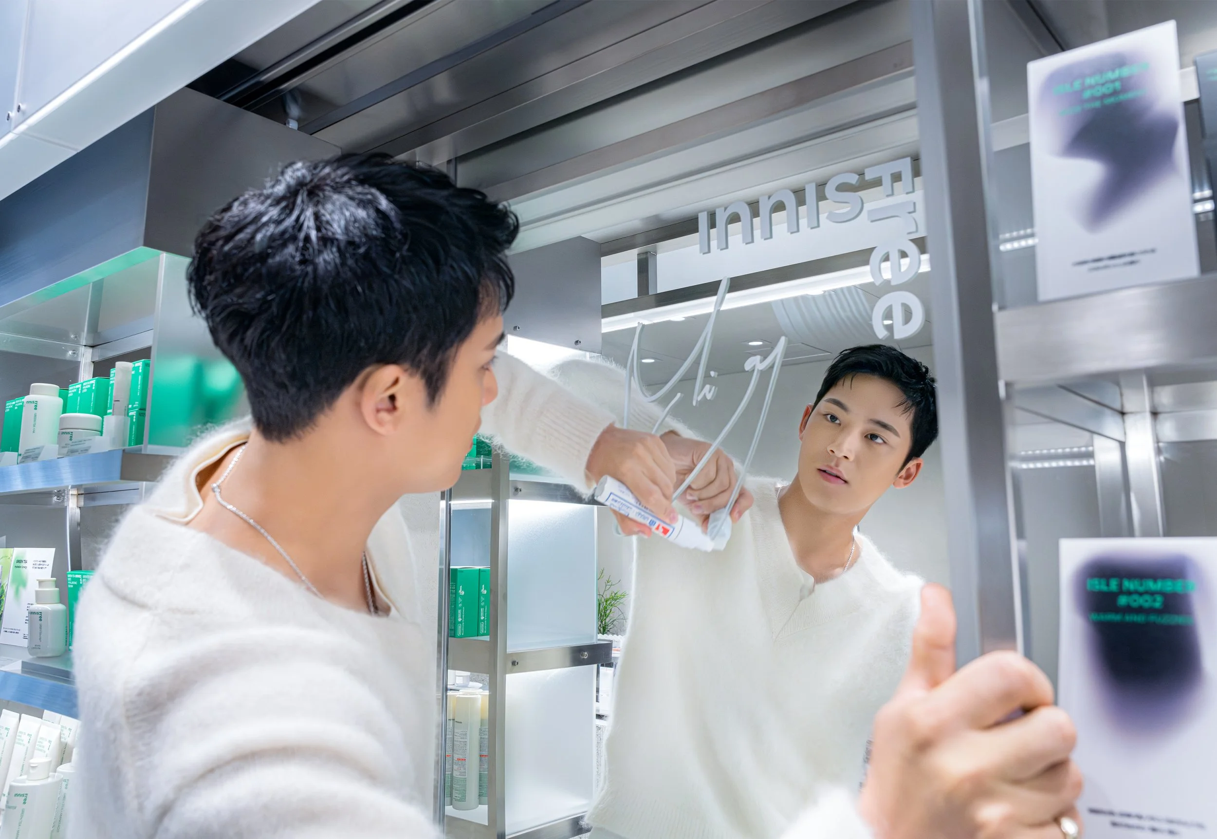 Two young men in cream sweaters looking at their reflections in a mirror inside a store, with one writing on the mirror with a marker, surrounded by shelves of skincare products.