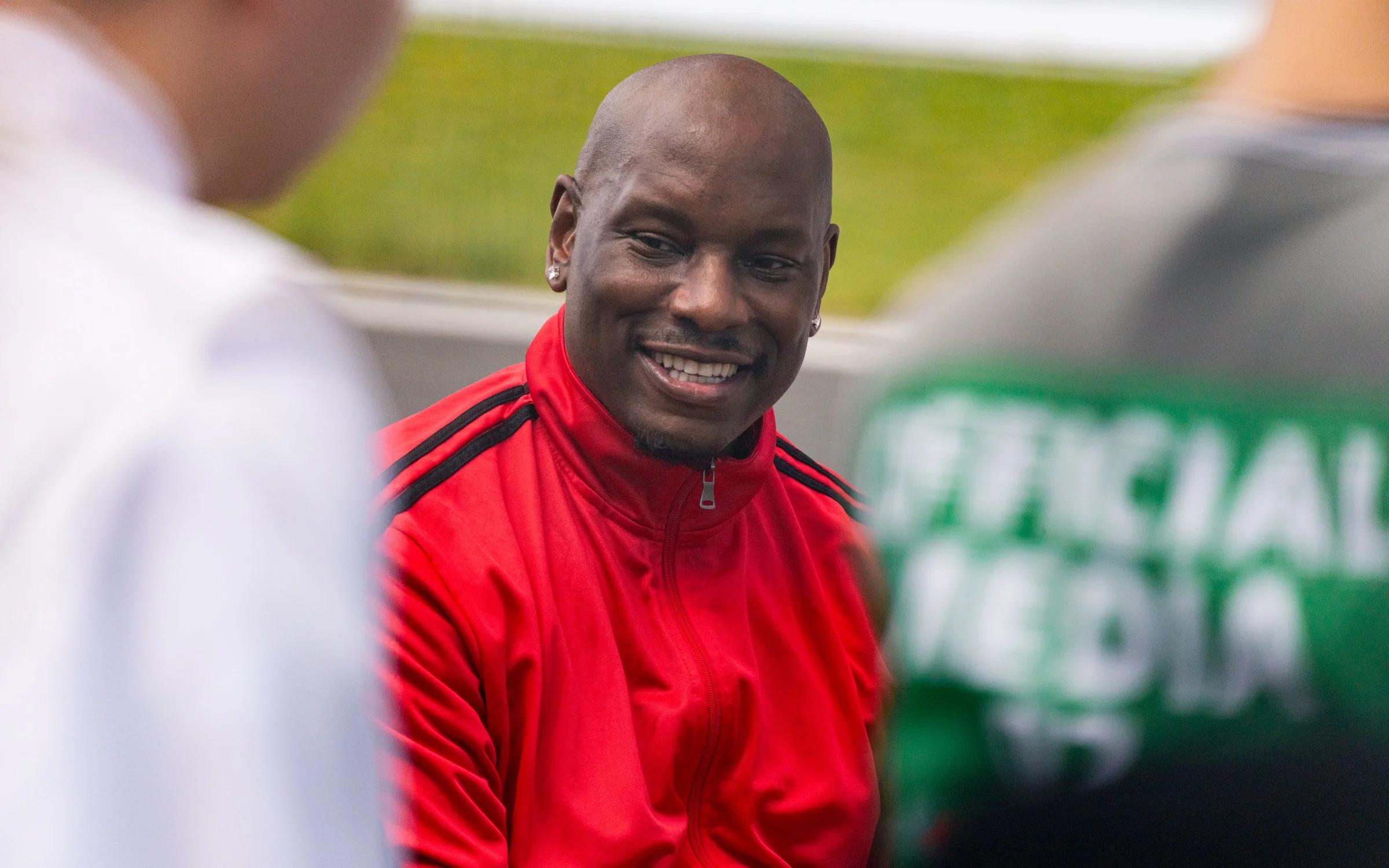 A smiling man wearing a red sports jacket with black stripes engaged in conversation with two other people, one in a white shirt and the other in a gray shirt with green lettering, outdoors on a sports field.