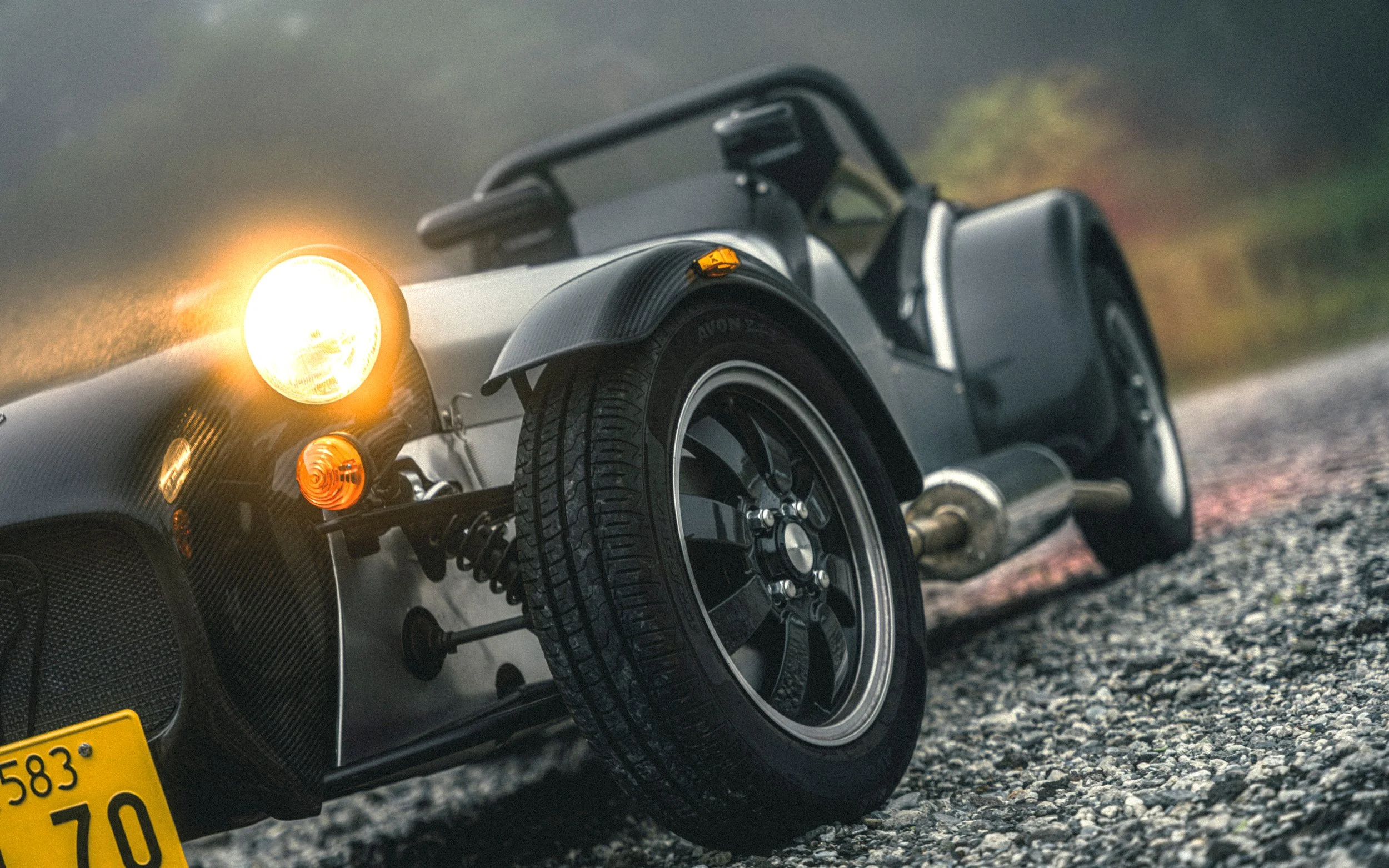 Close-up of a vintage three-wheeled sports car on a gravel road at sunset, showing the front tires, headlights, and part of the engine.