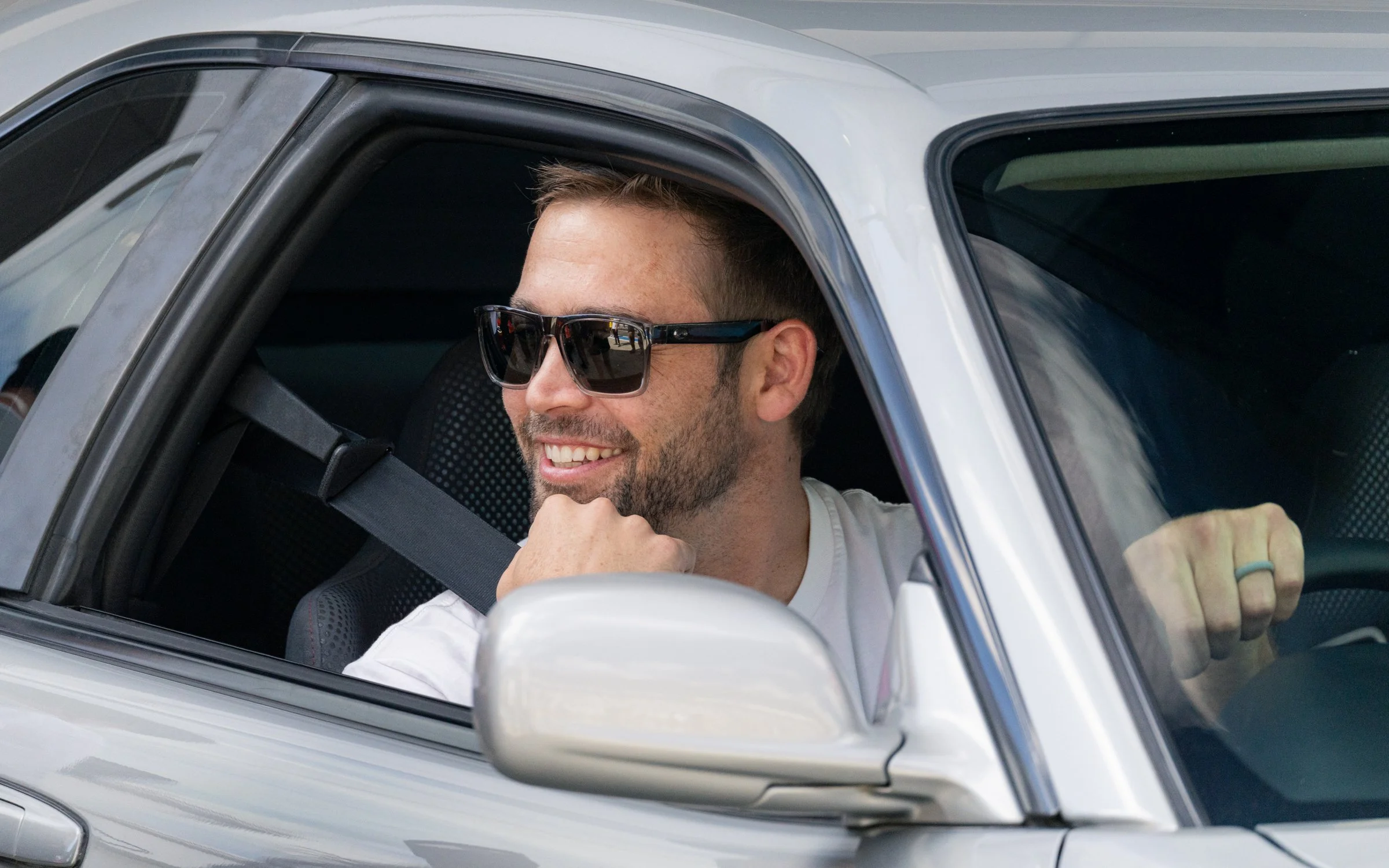 Smiling man with sunglasses sitting in the driver seat of a silver car, looking out the window.