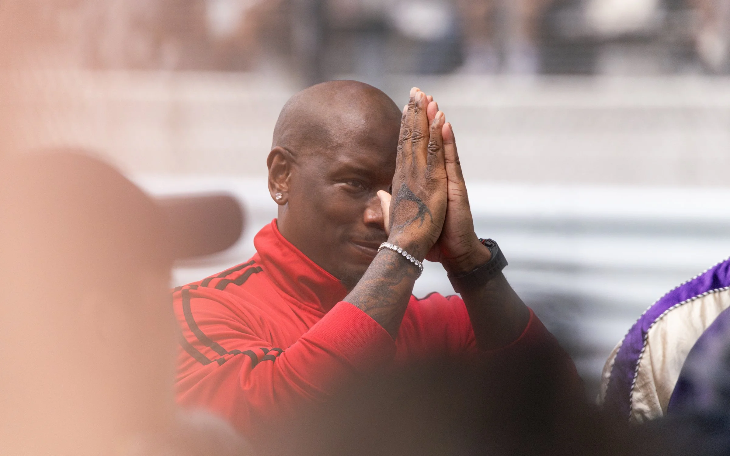 A man with a shaved head wearing a red sports jacket with black stripes, holding his hands together in a prayer position near his face, appears to be in a moment of reflection or prayer.