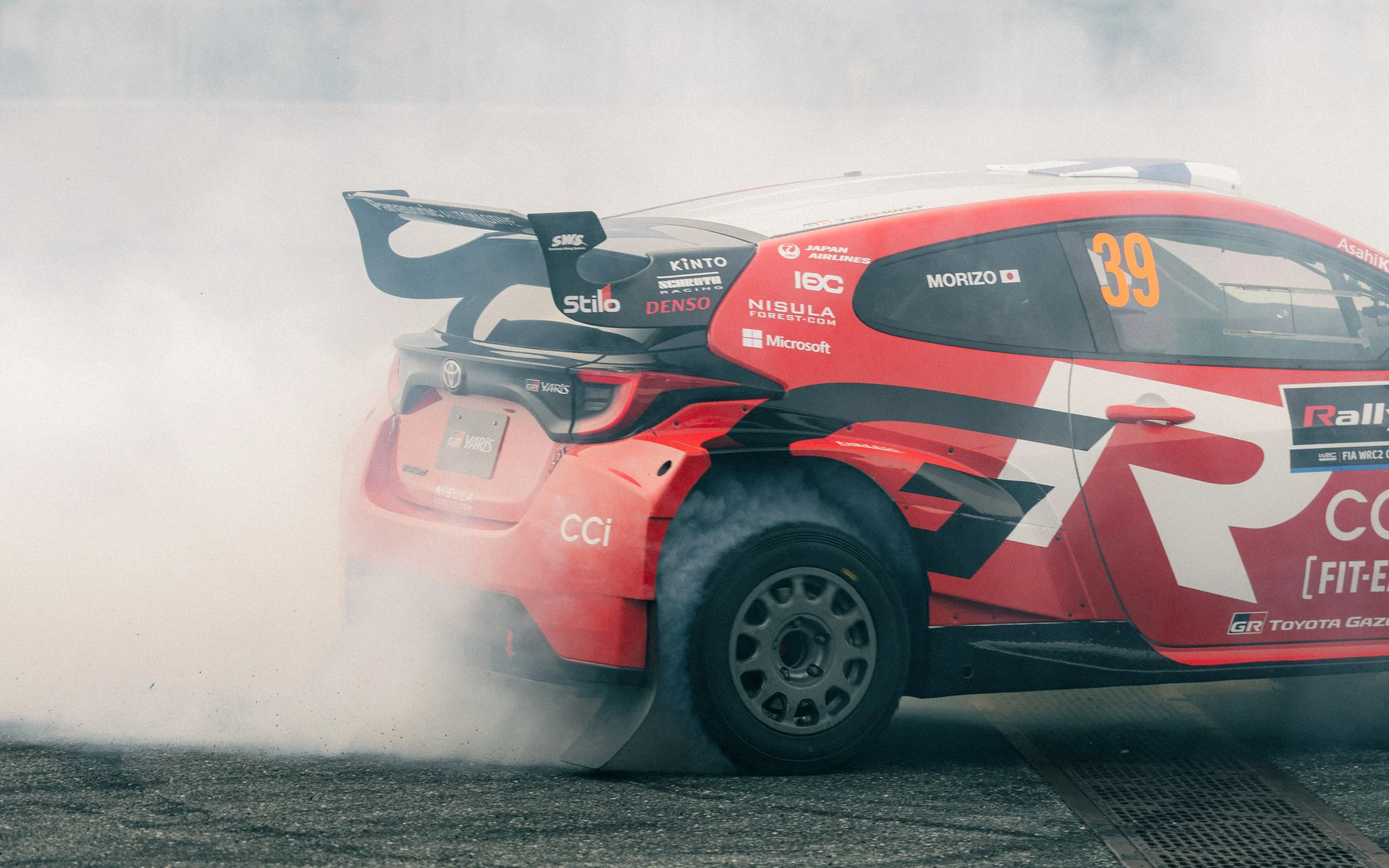 A red and black Toyota rally car with the number 39 on the side, emitting smoke from the rear tires during a race.