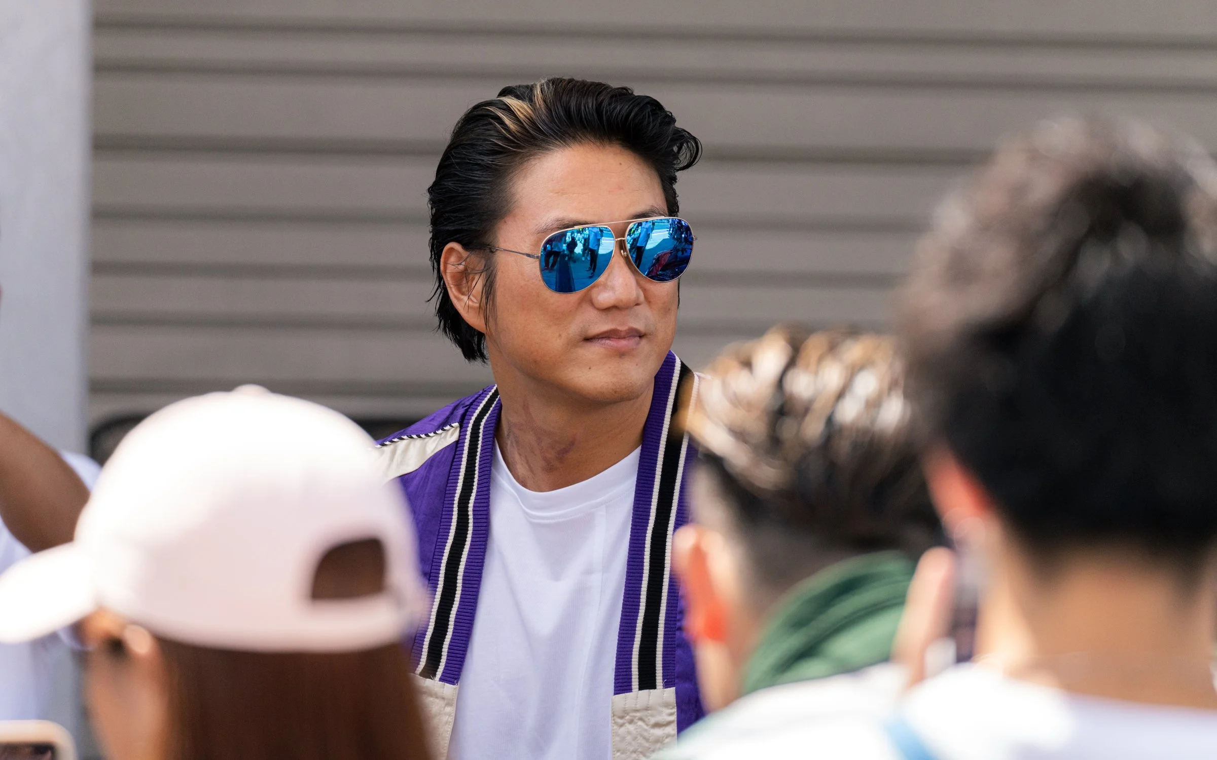 A man wearing reflective blue sunglasses and a purple jacket with white stripes, standing among a crowd.