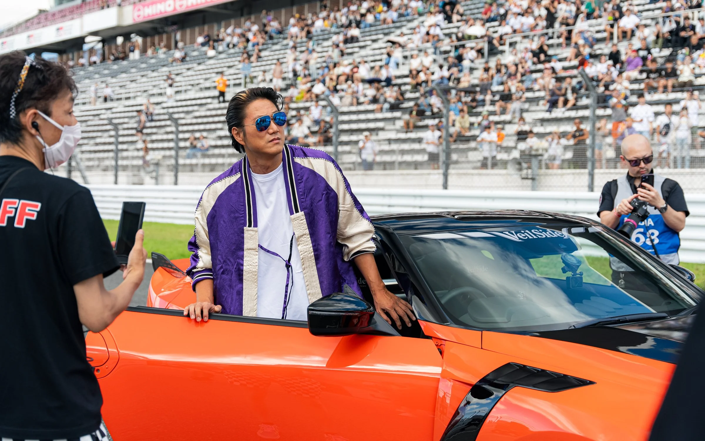 A man with sunglasses and a purple and beige jacket leaning on an orange sports car at a racetrack, with a crowd in the stands and two photographers nearby.