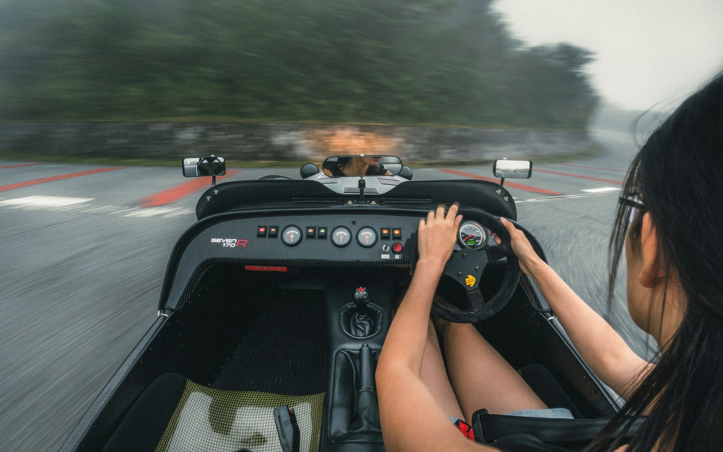 A person driving a race car on a wet track, with trees blurred in the background.