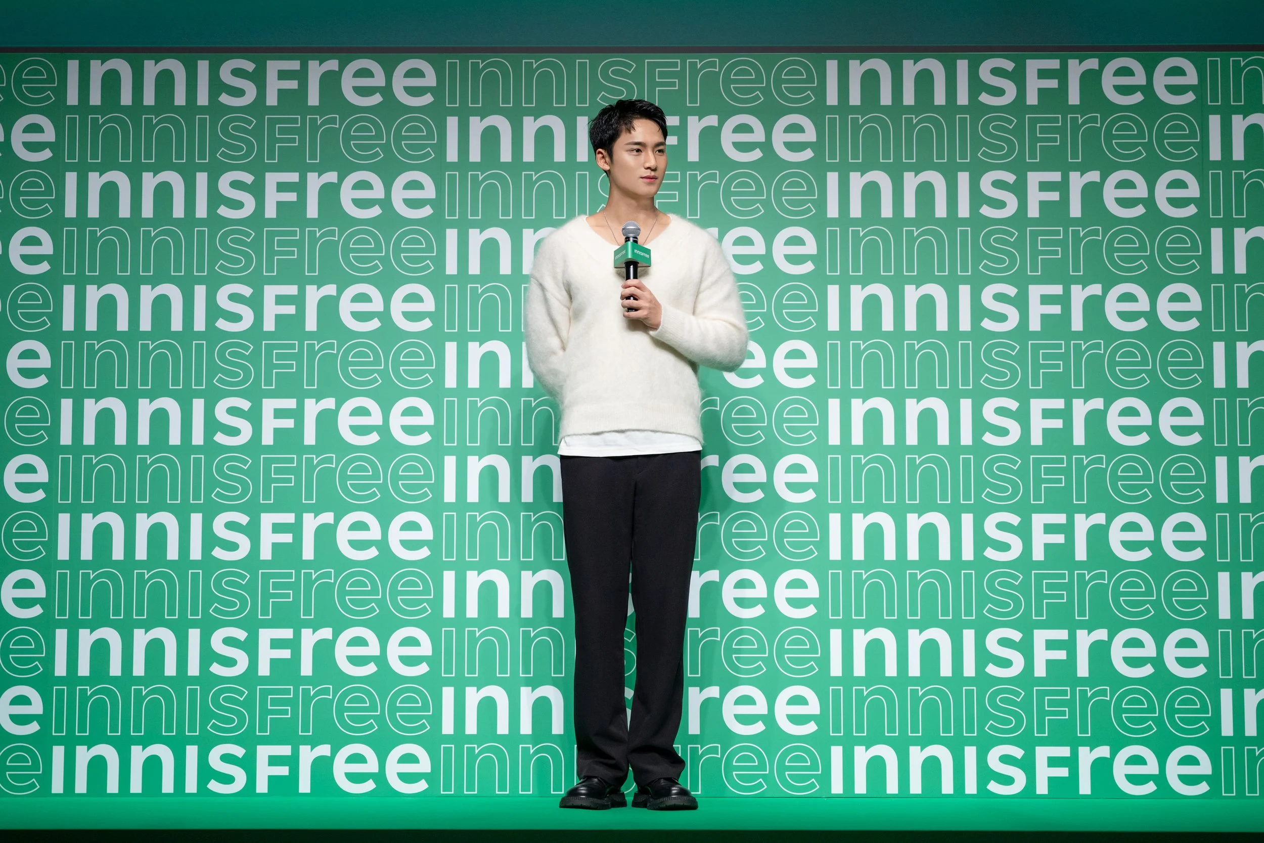 A young man standing on a green stage backdrop with the word 'innisfree' repeatedly printed in white and green. He is holding a microphone and looking slightly to his right, wearing a cream-colored sweater, black pants, and black shoes.
