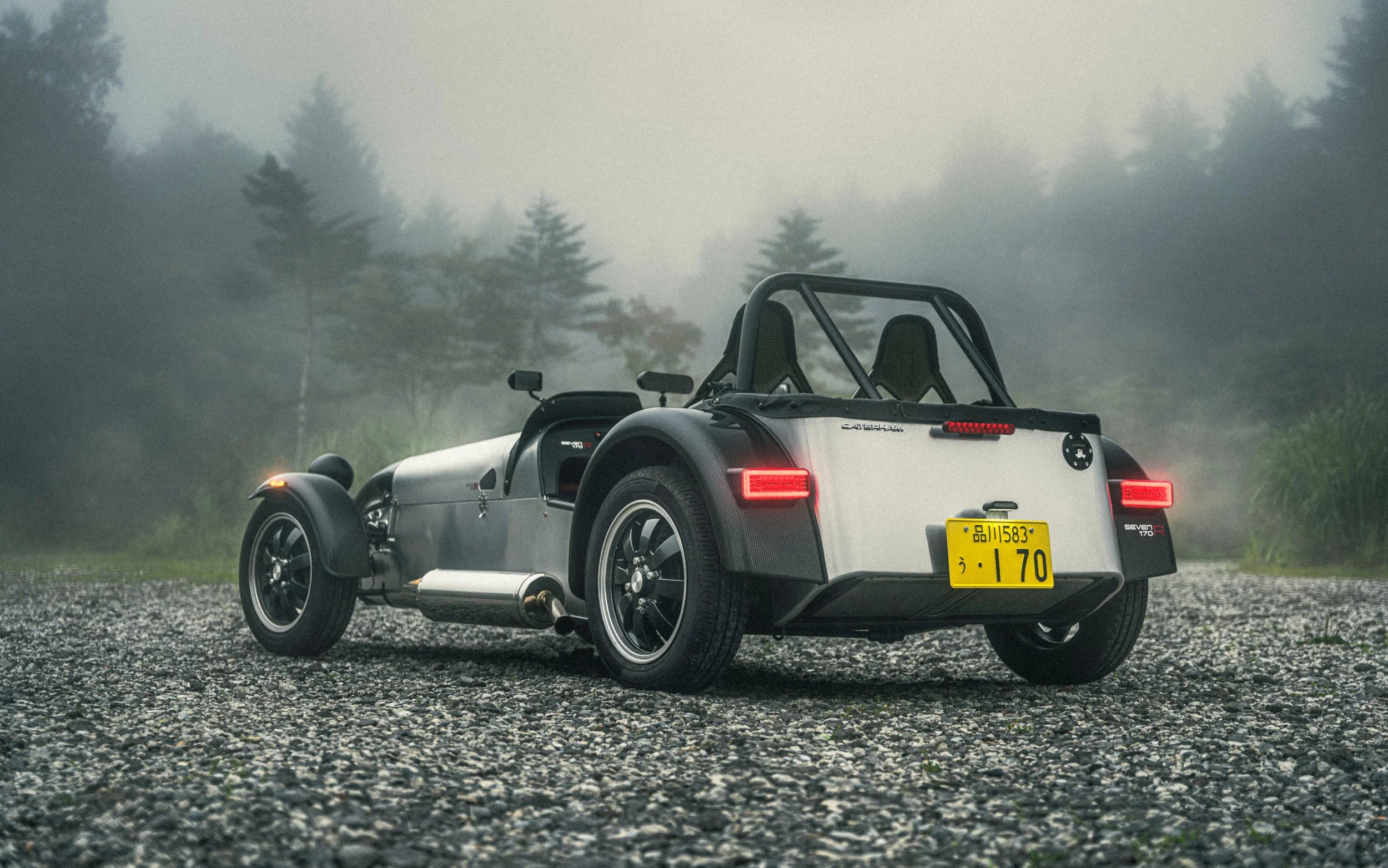 A two-seater vintage-style sports car with a roll cage parked on a gravel surface in a foggy, wooded area.
