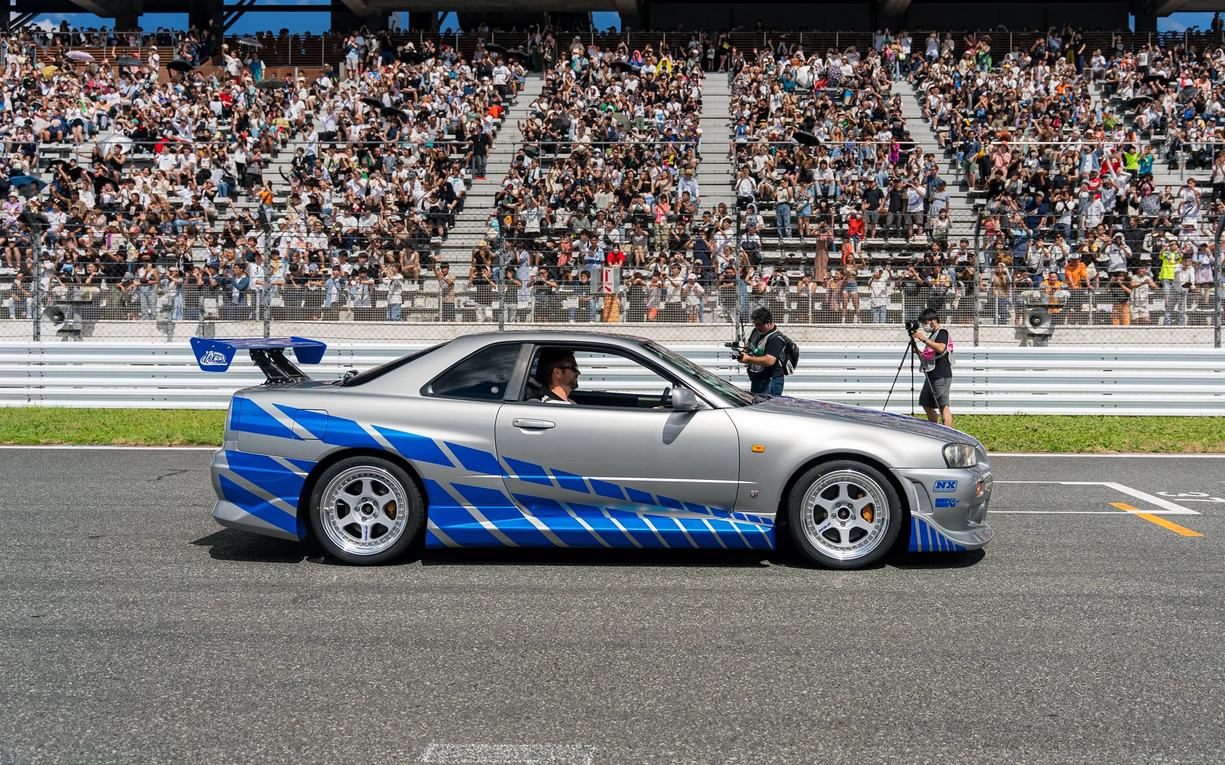 A silver race car with blue and white decals on a race track, with a driver inside, and a large crowd in the grandstands in the background. Photographers are capturing the moment.