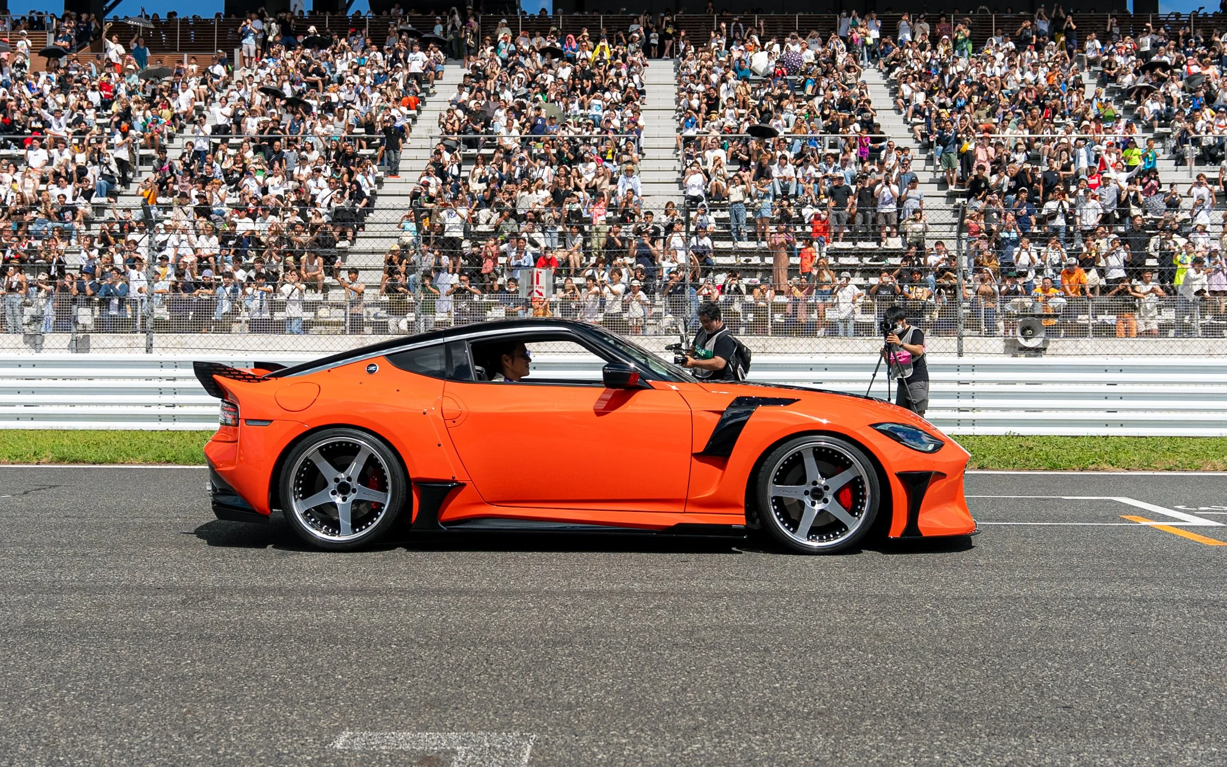 A bright orange sports car is parked at a race track, with a large crowd of spectators in the grandstands behind a fence, and two photographers taking pictures of the car.