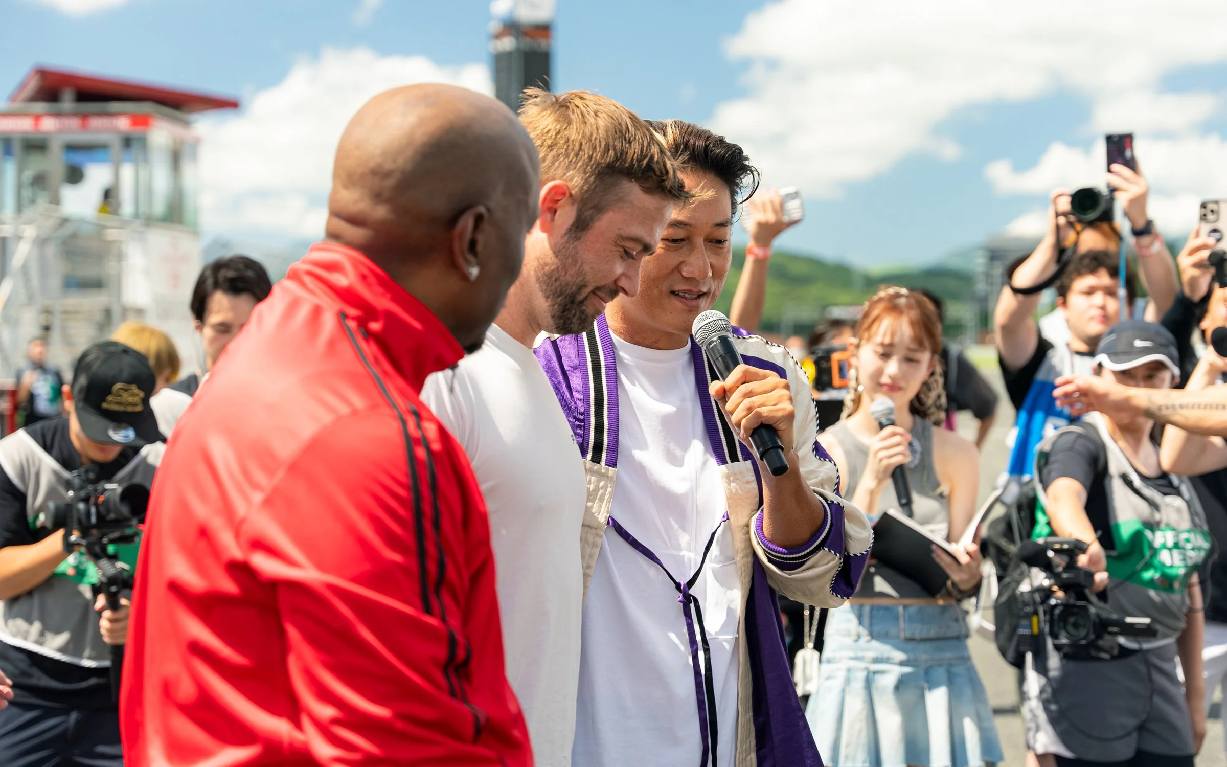 Three men are standing together outside on a sunny day, with the man on the right speaking into a microphone. They are surrounded by a crowd of photographers and reporters, some taking pictures and others holding microphones.