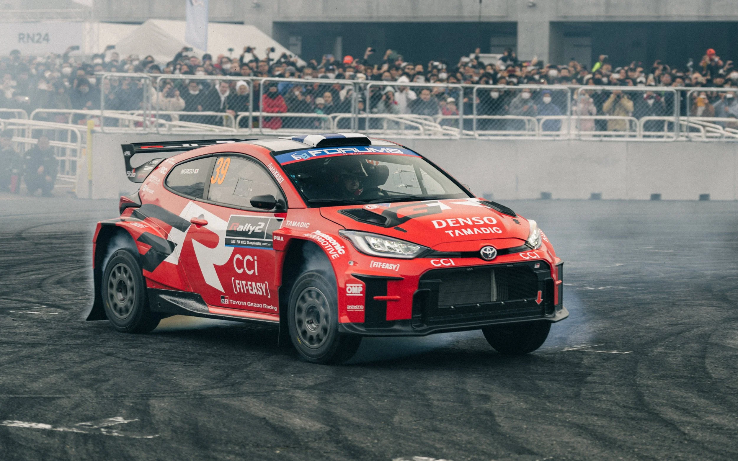A red Toyota Gazoo Racing rally car with the number 39 on the side, drifting on a race track with smoke and tire marks, while a crowd of spectators watches from the stands in the background.