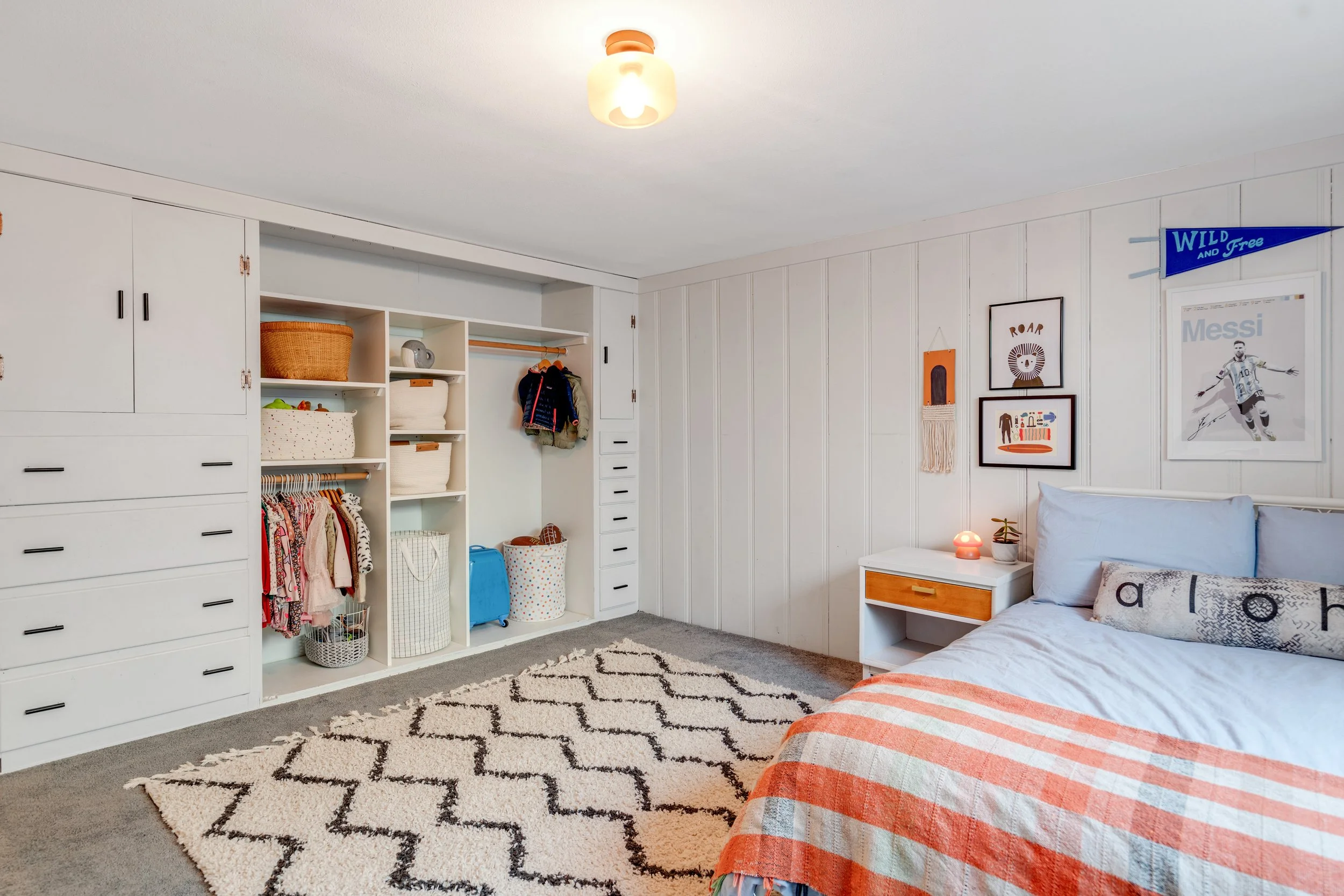 Bedroom with white walls, a bed with light blue bedding and an orange striped blanket, a nightstand with a lamp and decor, framed art on the wall, a closet with open shelves and hanging clothes, a black and white patterned rug, and a ceiling light.