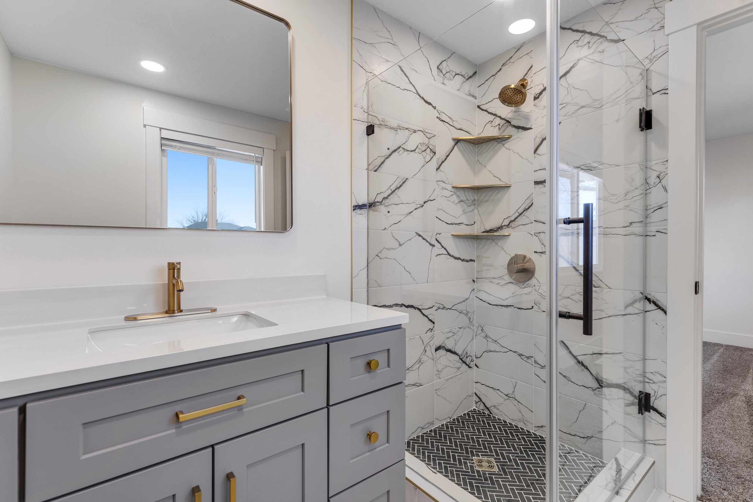 Modern bathroom with gray vanity, white countertop, large mirror, and a walk-in shower with marble tiles, gold fixtures, and black patterned floor tiles.