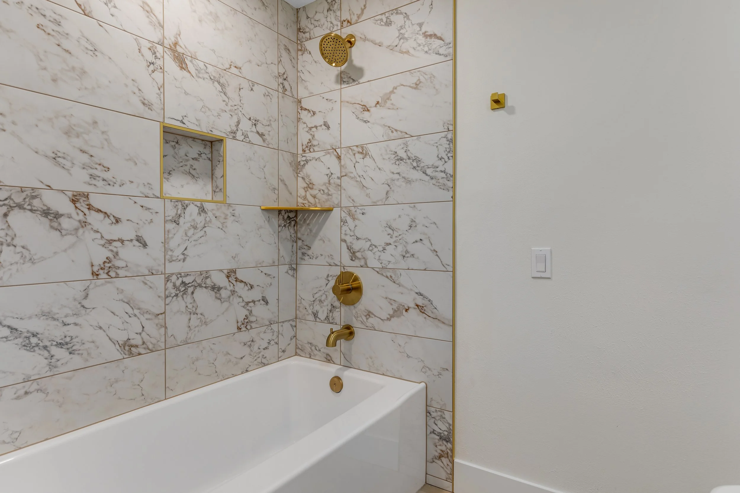 Bathroom with marble tiles, gold fixtures, and a white bathtub.