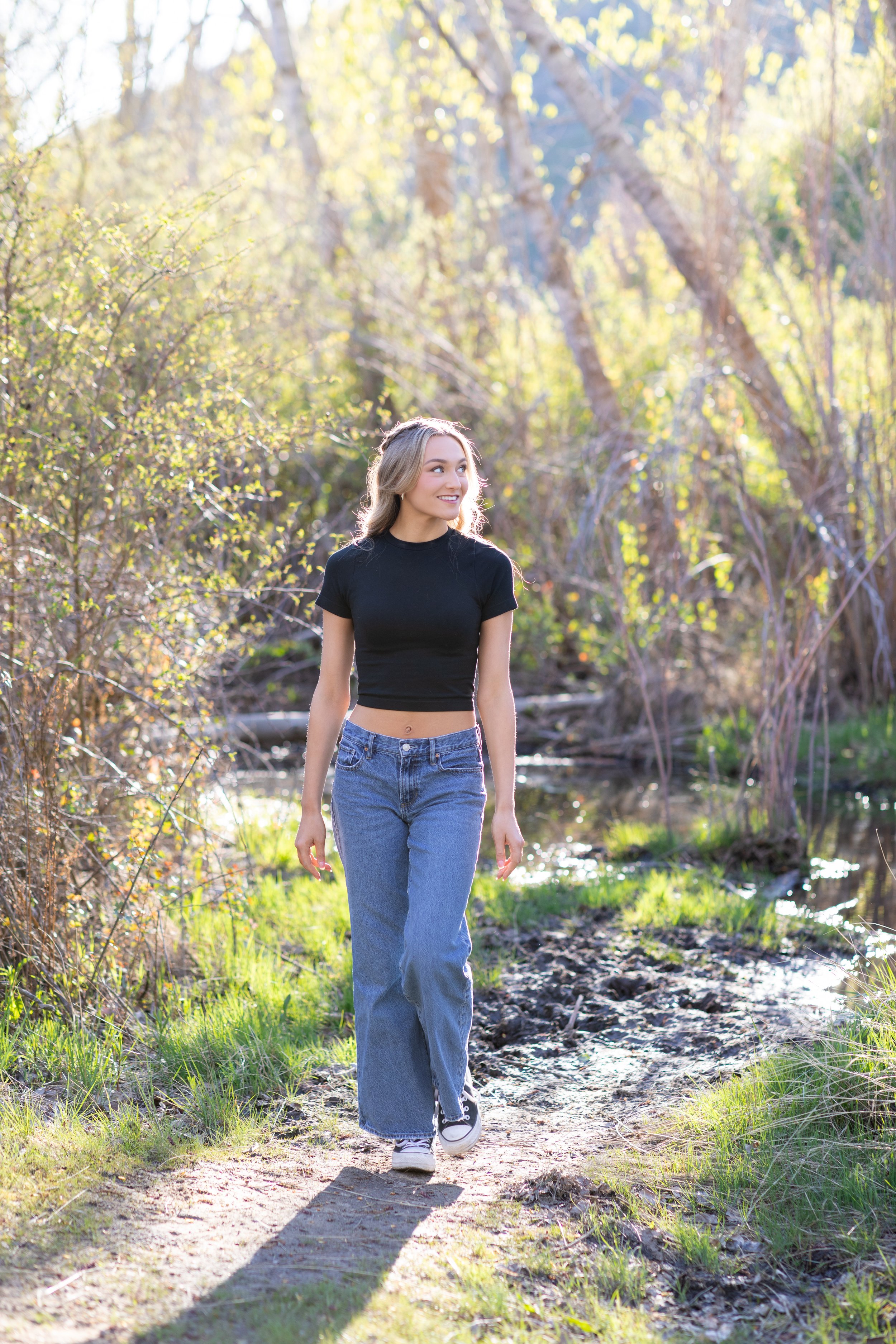 A young woman with long, wavy blonde hair, wearing a black crop top and blue jeans, walking on a dirt trail near a small creek surrounded by trees and greenery on a sunny day.
