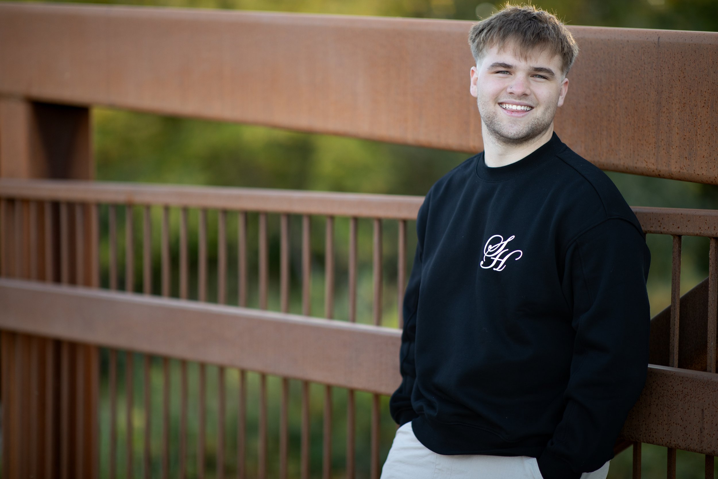 A young man with short, light brown hair and a beard, smiling and leaning against a wooden railing outdoors with a blurred green background, wearing a black sweatshirt with white embroidered initials 'H' on the chest.
