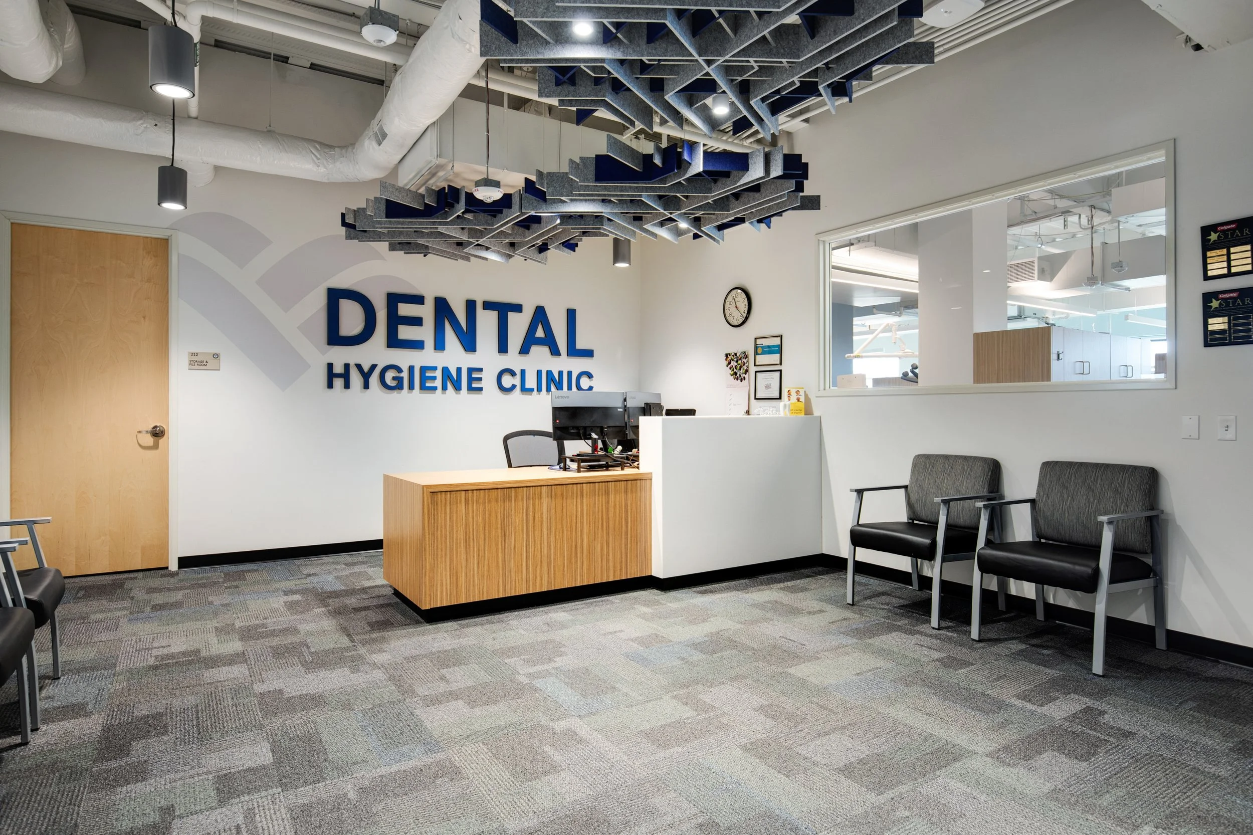 Reception area of a dental hygiene clinic with a wooden desk, computer, and two chairs along the wall. The wall has large blue letters that read 'DENTAL HYGIENE CLINIC', and a window looks into a brighter room with dental equipment.