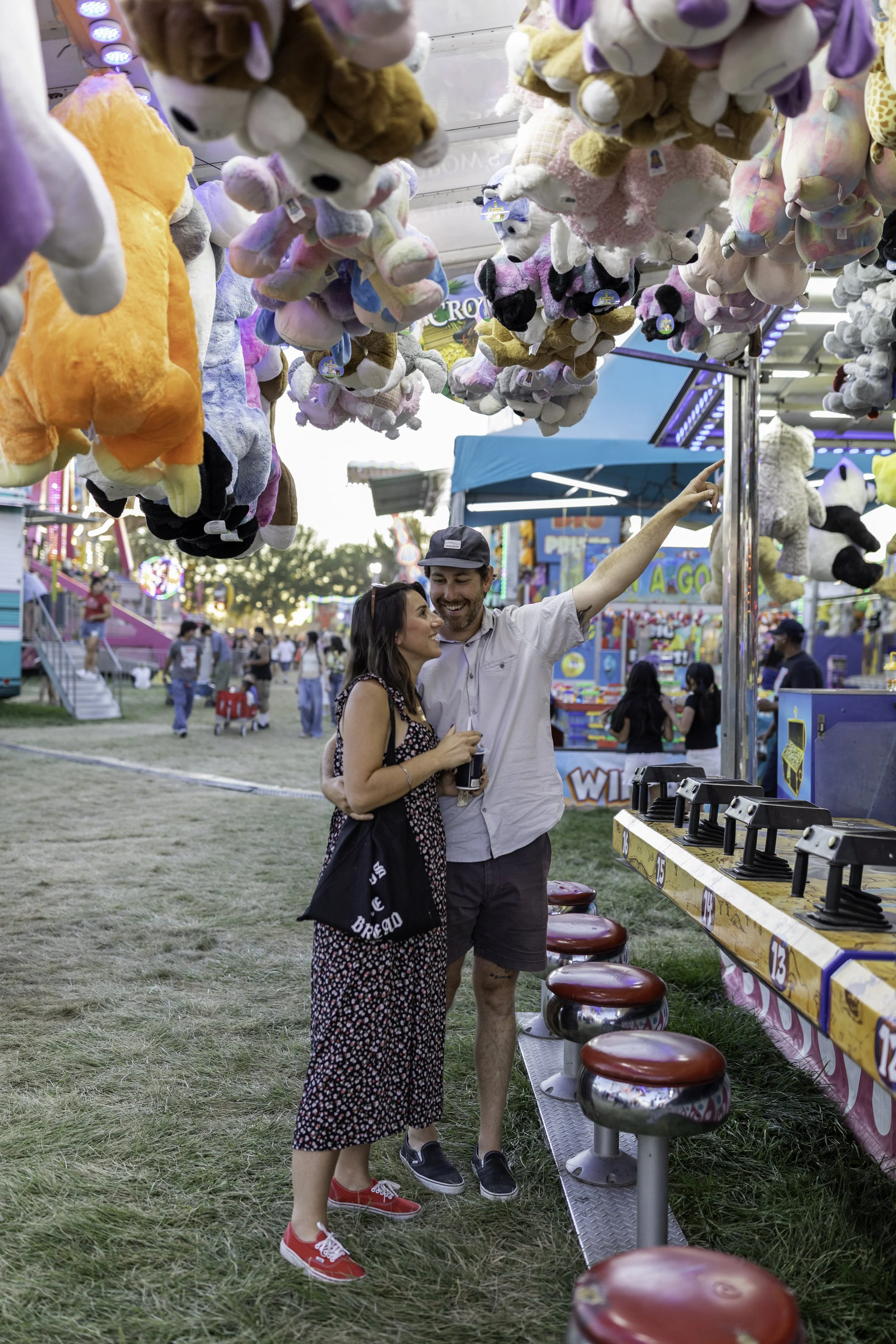 A couple at a carnival game booth, with plush stuffed animals hanging above them and colorful booths in the background. The man is pointing at the stuffed animals while the woman looks on smiling.