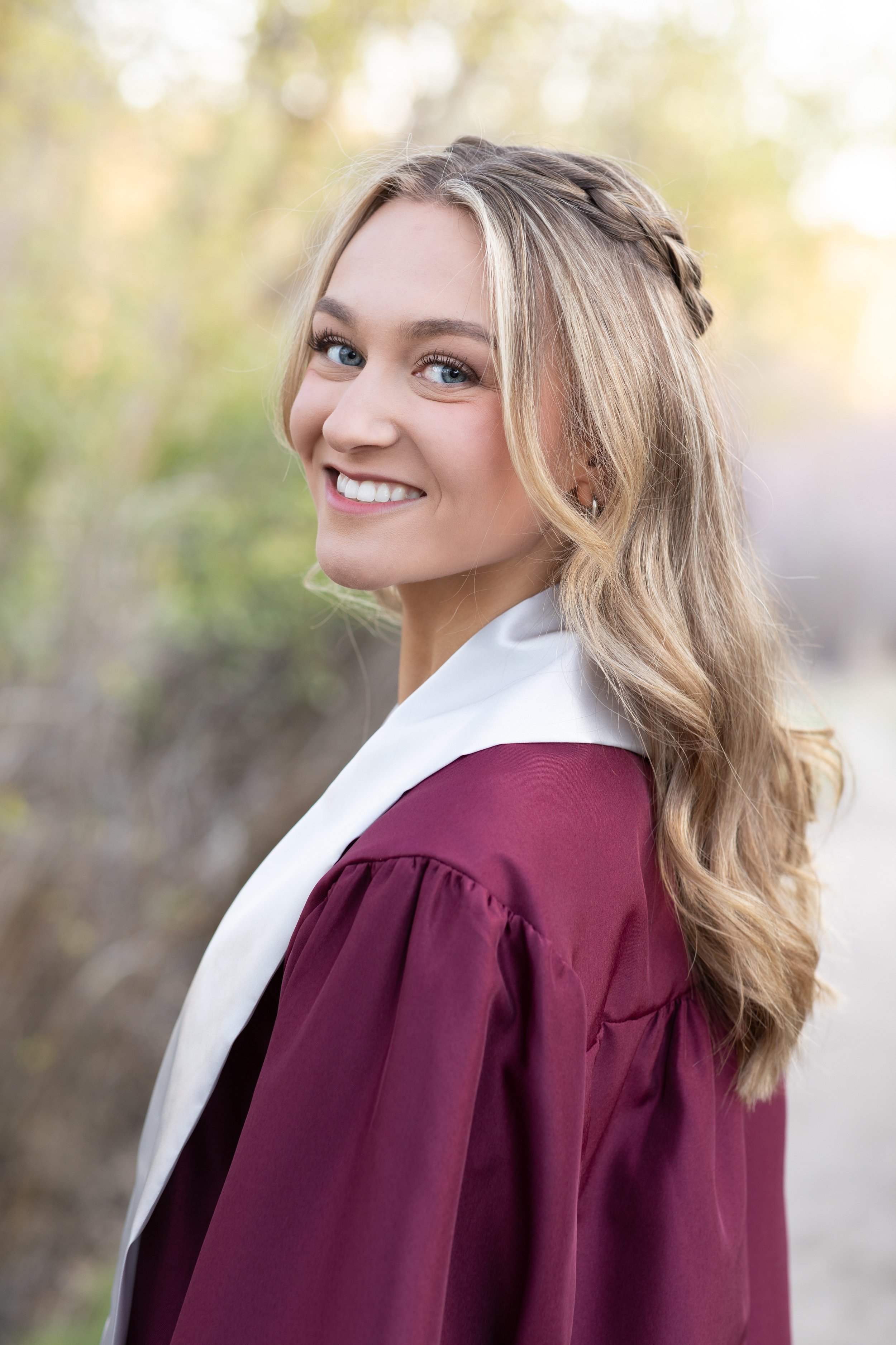 A young woman in a graduation gown smiling outdoors with trees blurred in the background.