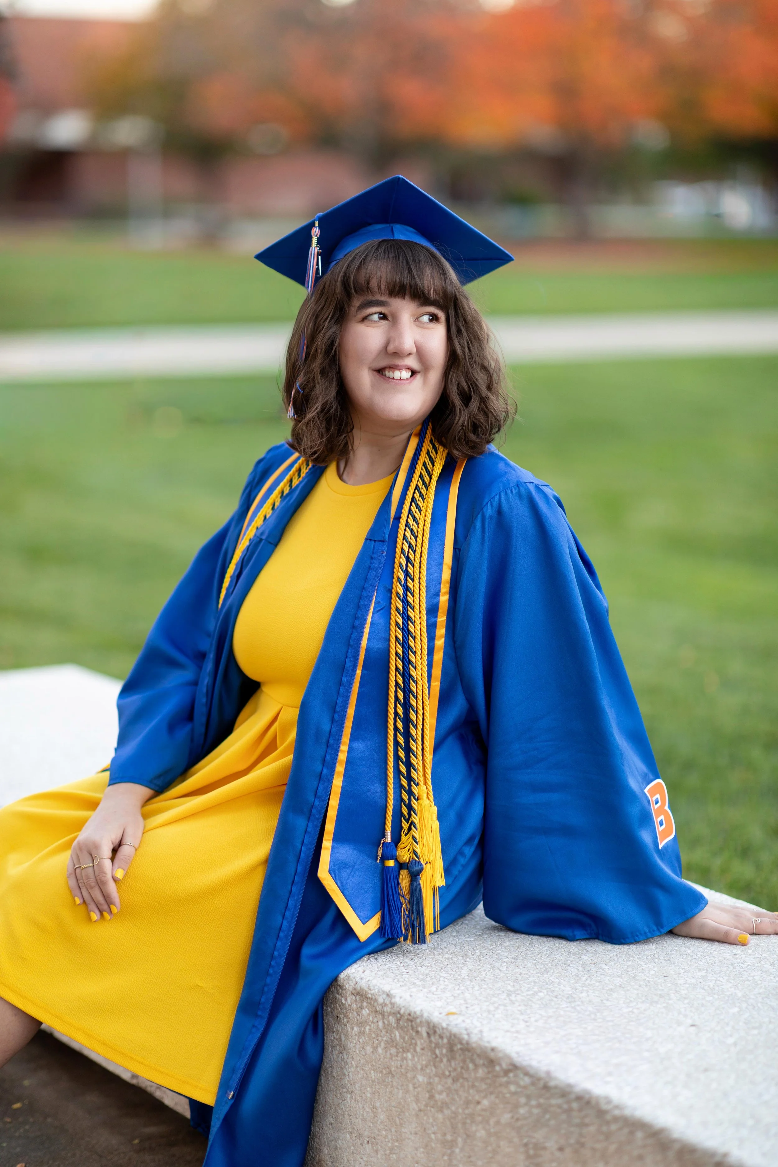 A young woman in a yellow dress and blue graduation gown and cap, sitting outdoors on a stone bench with a blurred background of grass and trees that have autumn-colored leaves.