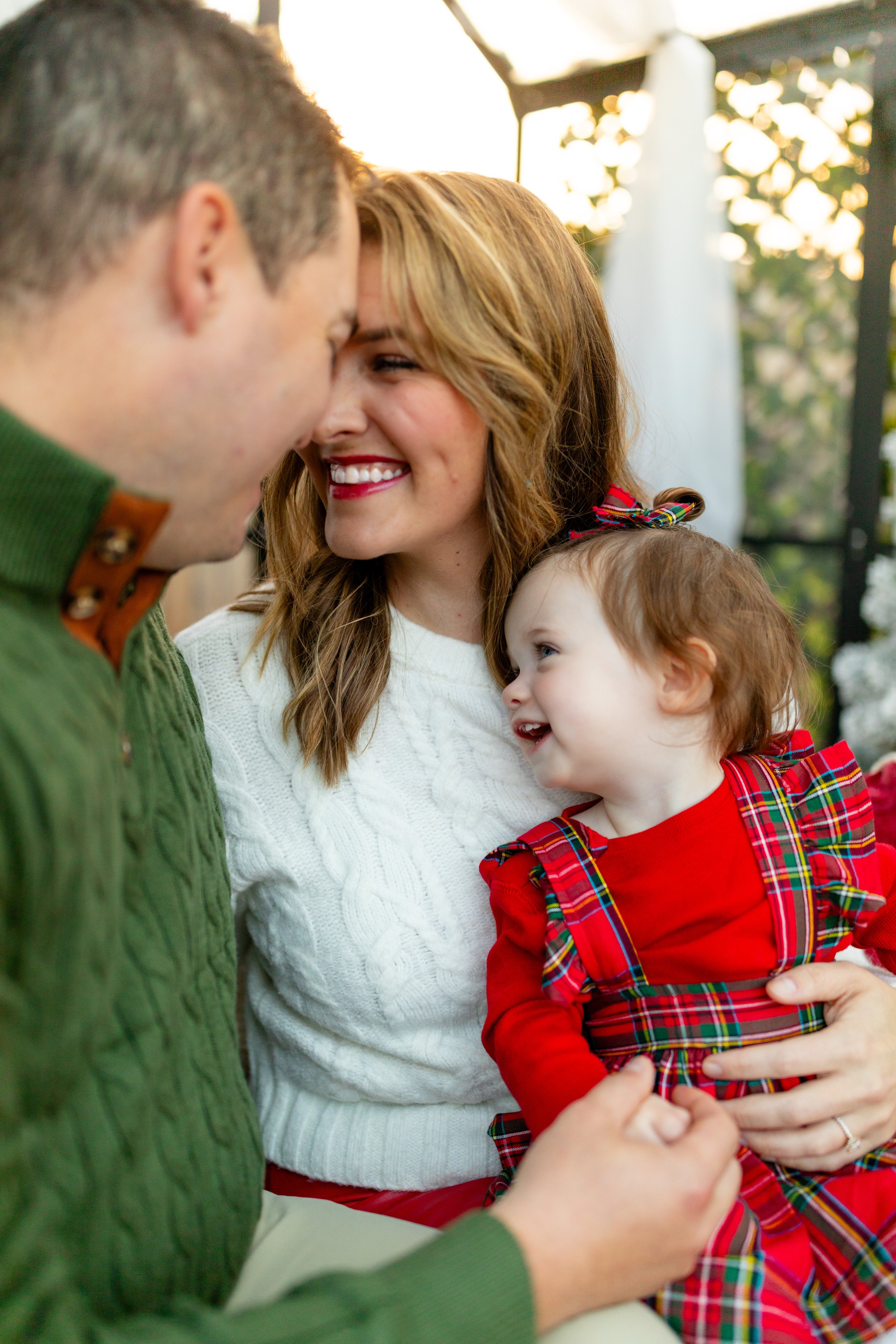 A smiling woman and a young girl with red hair and blue eyes, dressed in a red plaid dress, sharing a joyful moment as they sit close together outdoors in warm light, with a man partially visible smiling and holding the girl's hand.