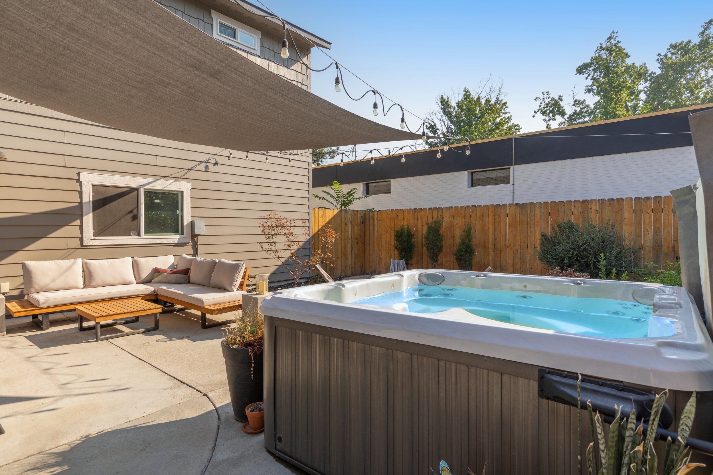 An outdoor patio with a hot tub, a beige sectional sofa with cushions, string lights overhead, potted plants, and a wooden privacy fence.