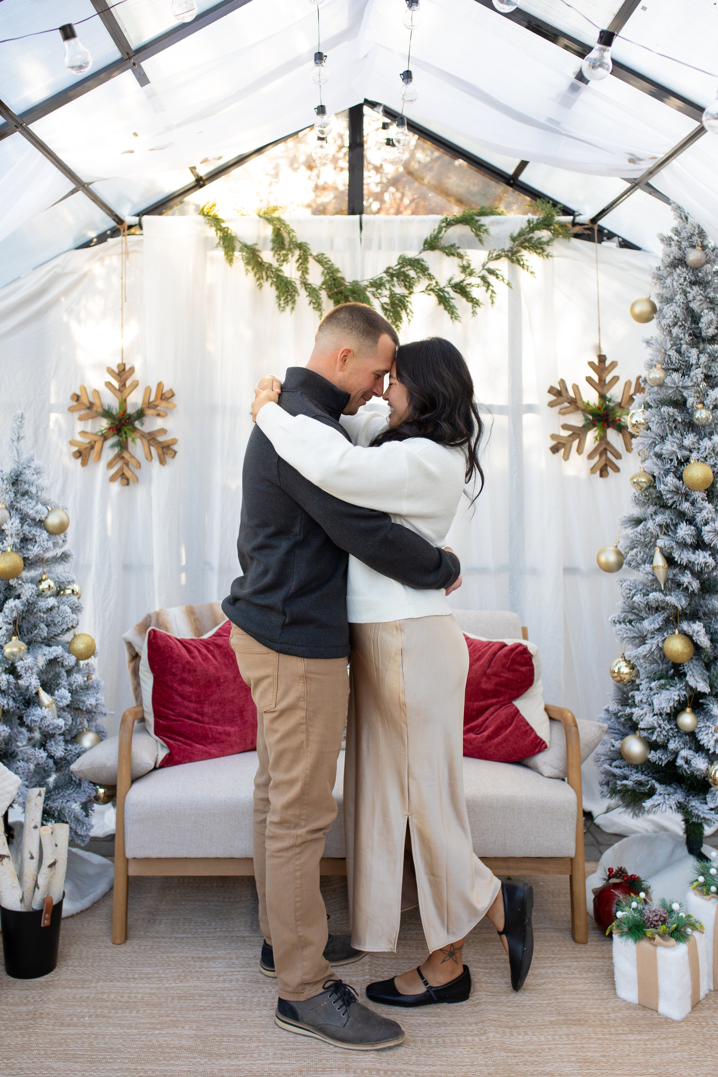 A couple embraces each other in a festive holiday setting with Christmas trees, snowflake ornaments, and wrapped gifts, inside a decorated tent.