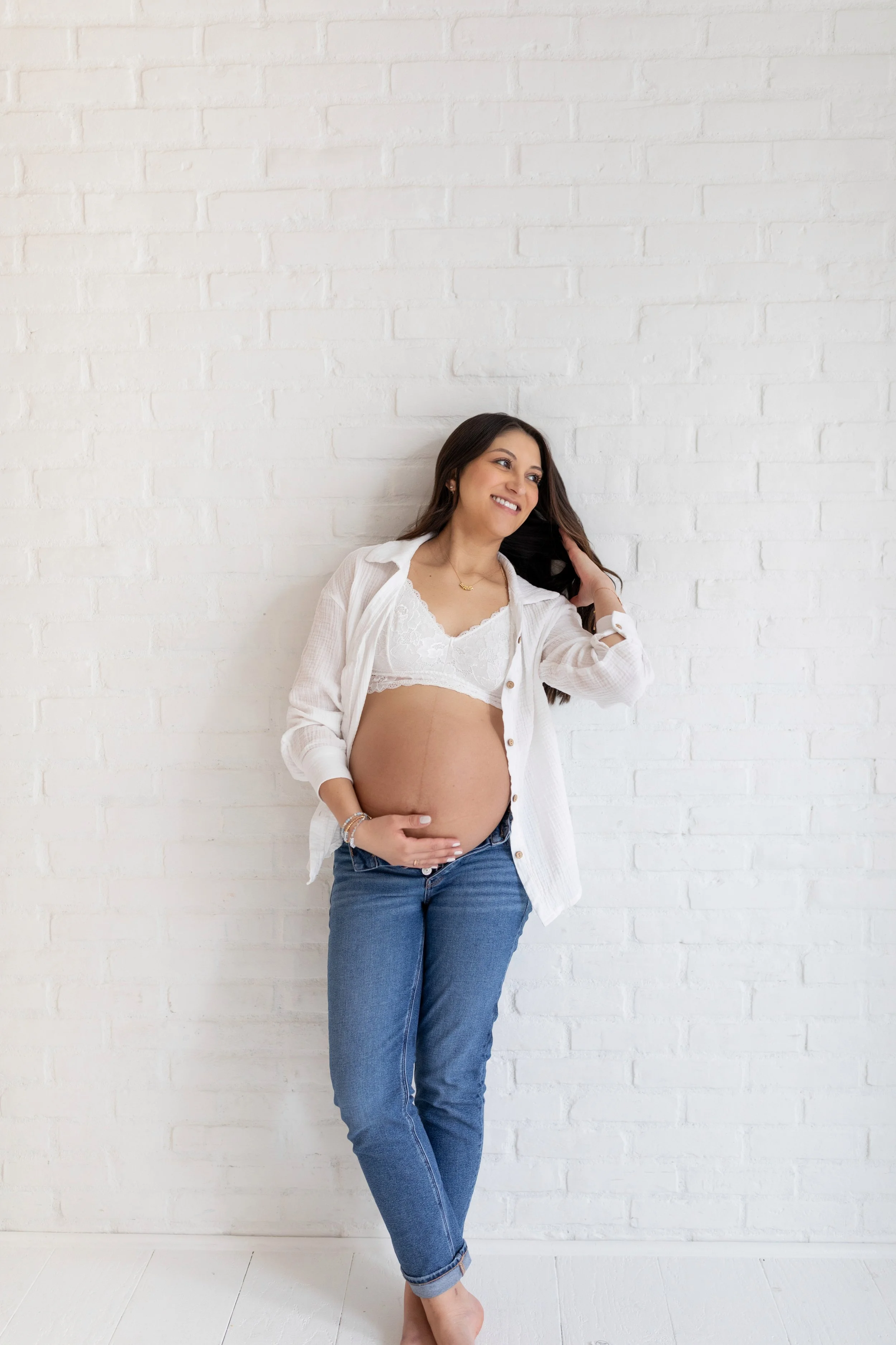 A pregnant woman with long dark hair smiling, wearing a white lace bra, open white shirt, and blue jeans, standing barefoot against a white brick wall.