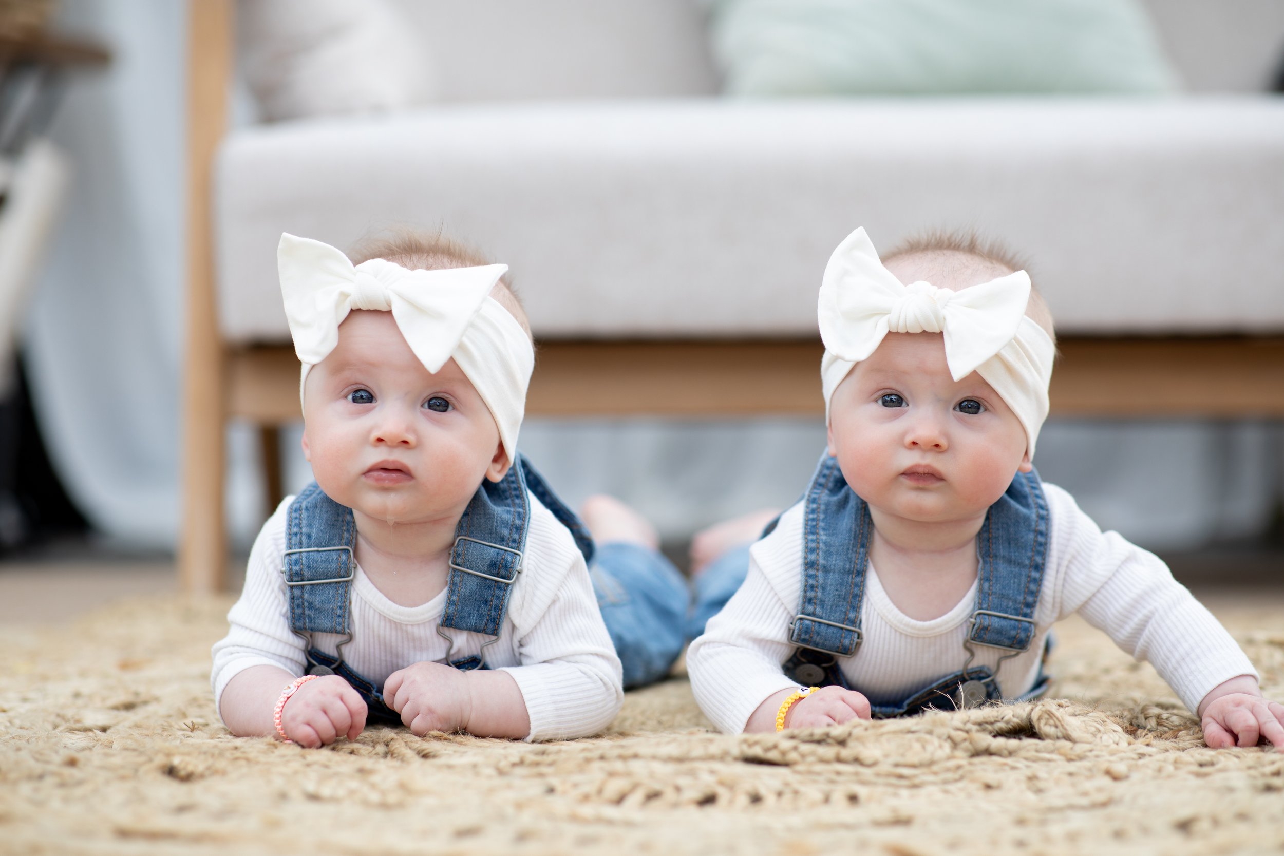 Two babies lying on their stomachs on a rug, wearing white headbands with large bows and denim overalls, indoors.