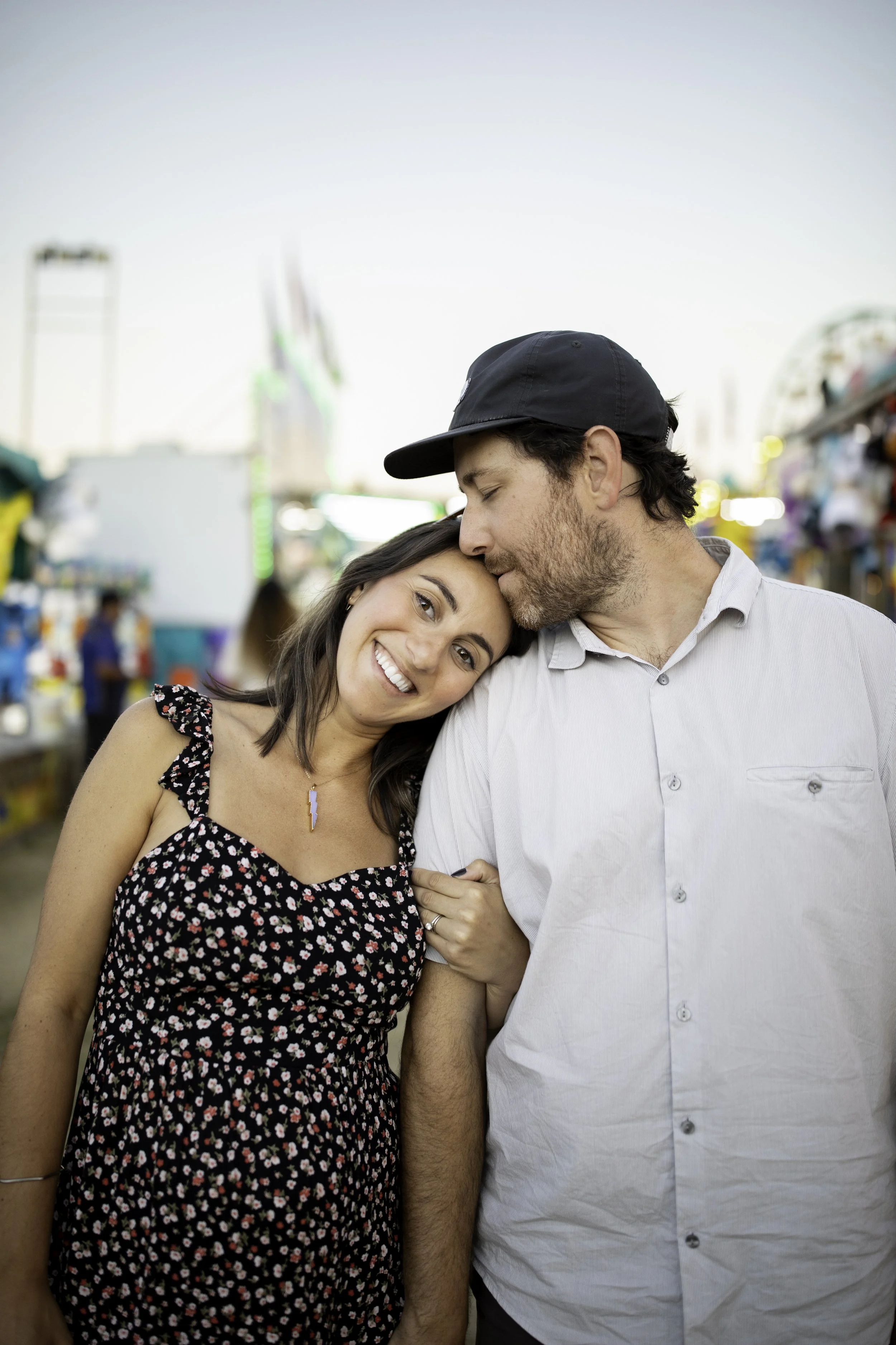 A smiling woman with shoulder-length dark hair wearing a floral dress and a necklace with a lightning bolt charm is leaning her head on the forehead of a man with a beard and a black cap, who is gently kissing her forehead. They are at an outdoor fai