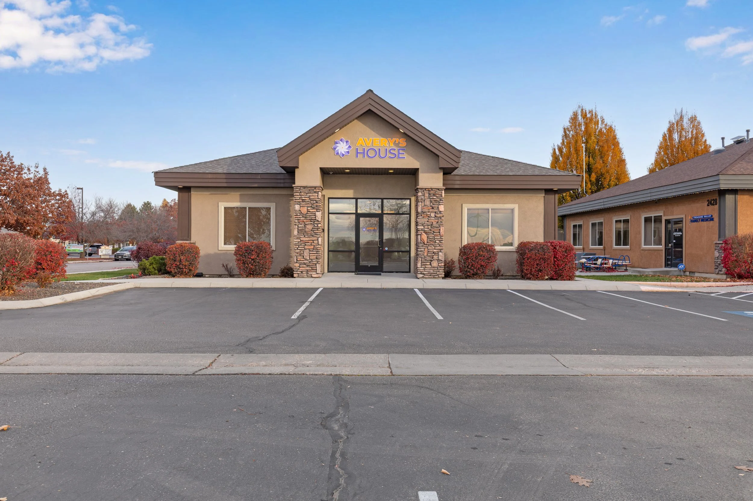 A beige commercial building with a sign that reads 'Avery's House' on the front, surrounded by red bushes and a parking lot with empty spaces, some trees with autumn foliage in the background.
