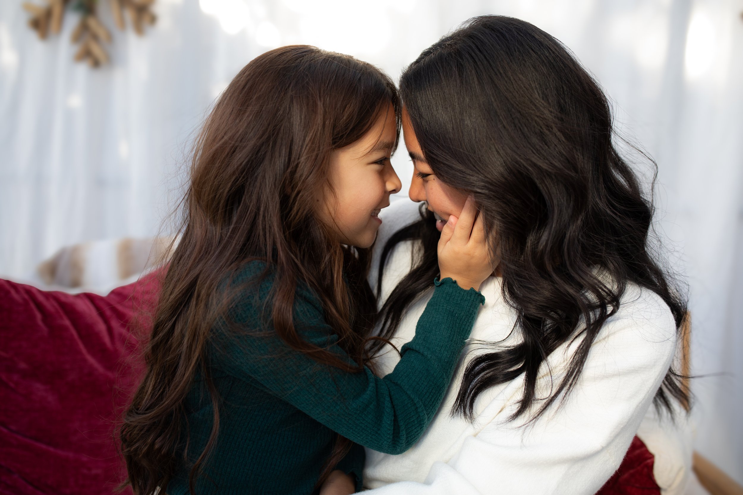 A woman and a girl with long brown hair sharing a close, smiling moment indoors, with a blurred white curtain background.