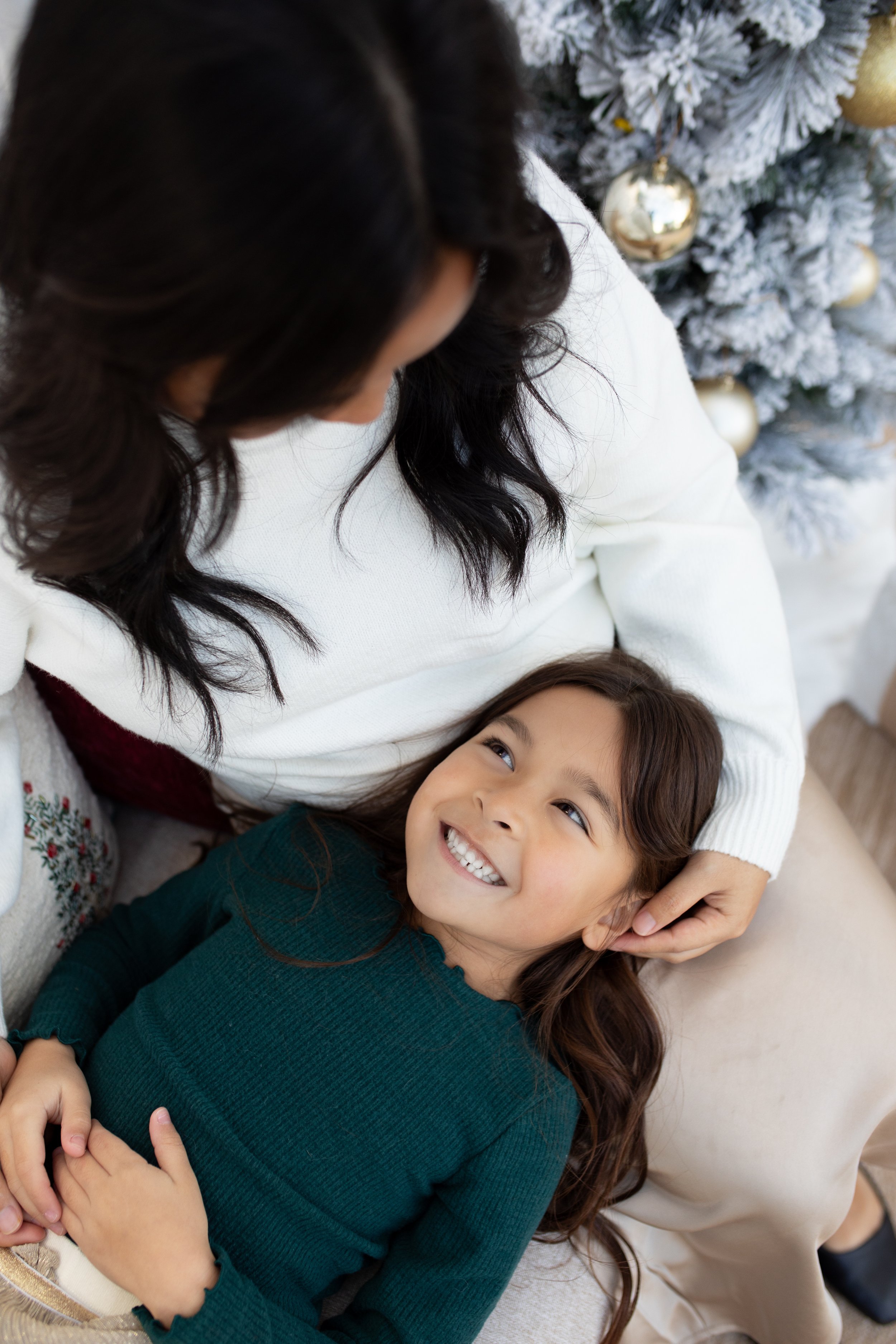 A woman and a young girl sitting together near a decorated Christmas tree, smiling and looking at each other.