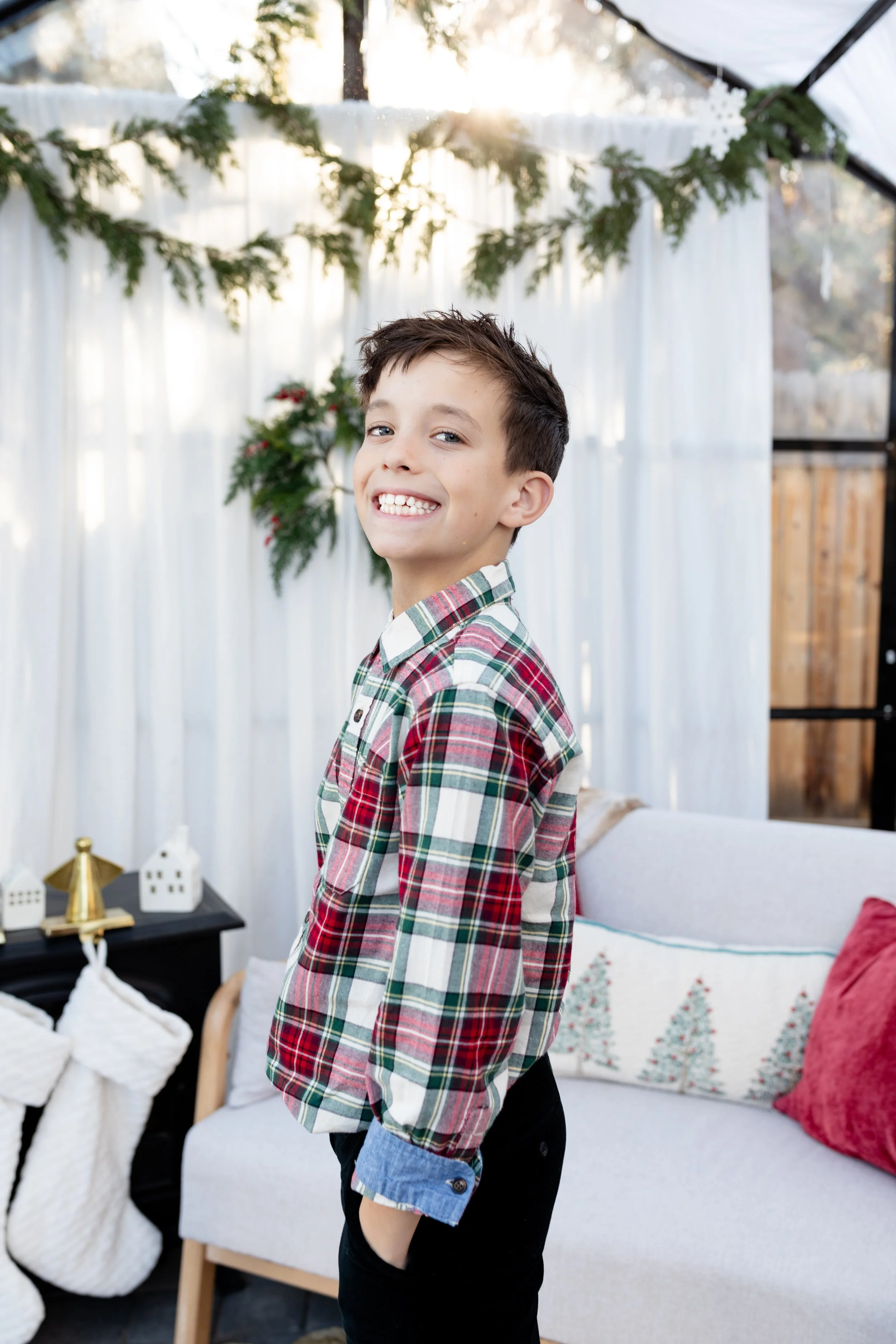 A smiling young boy with brown hair wearing a red, green, and white plaid shirt and dark pants standing indoors near a cozy decorated holiday setting.